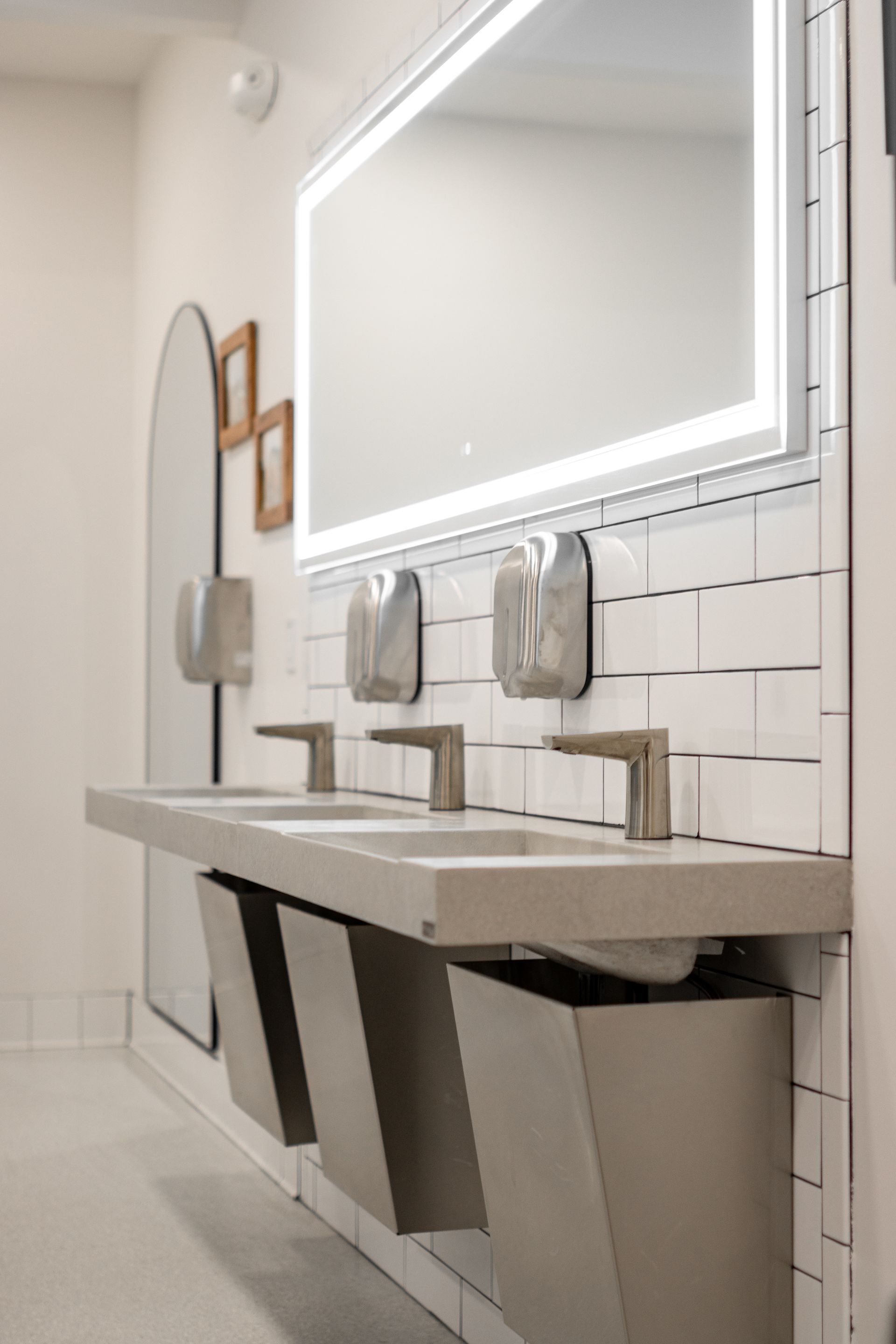 Modern restroom with illuminated mirror, sinks, and stalls in white and gray tiles