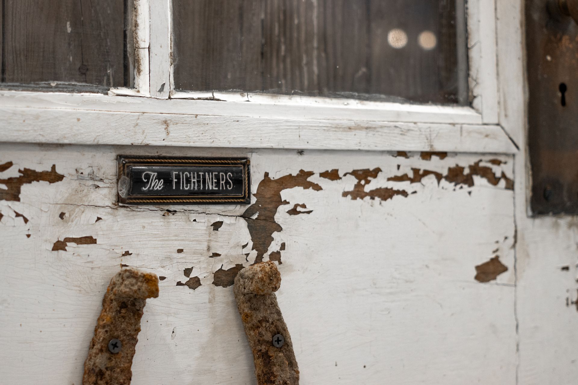 Weathered white door with peeling paint, small black plaque, and two rusty handles below a window.