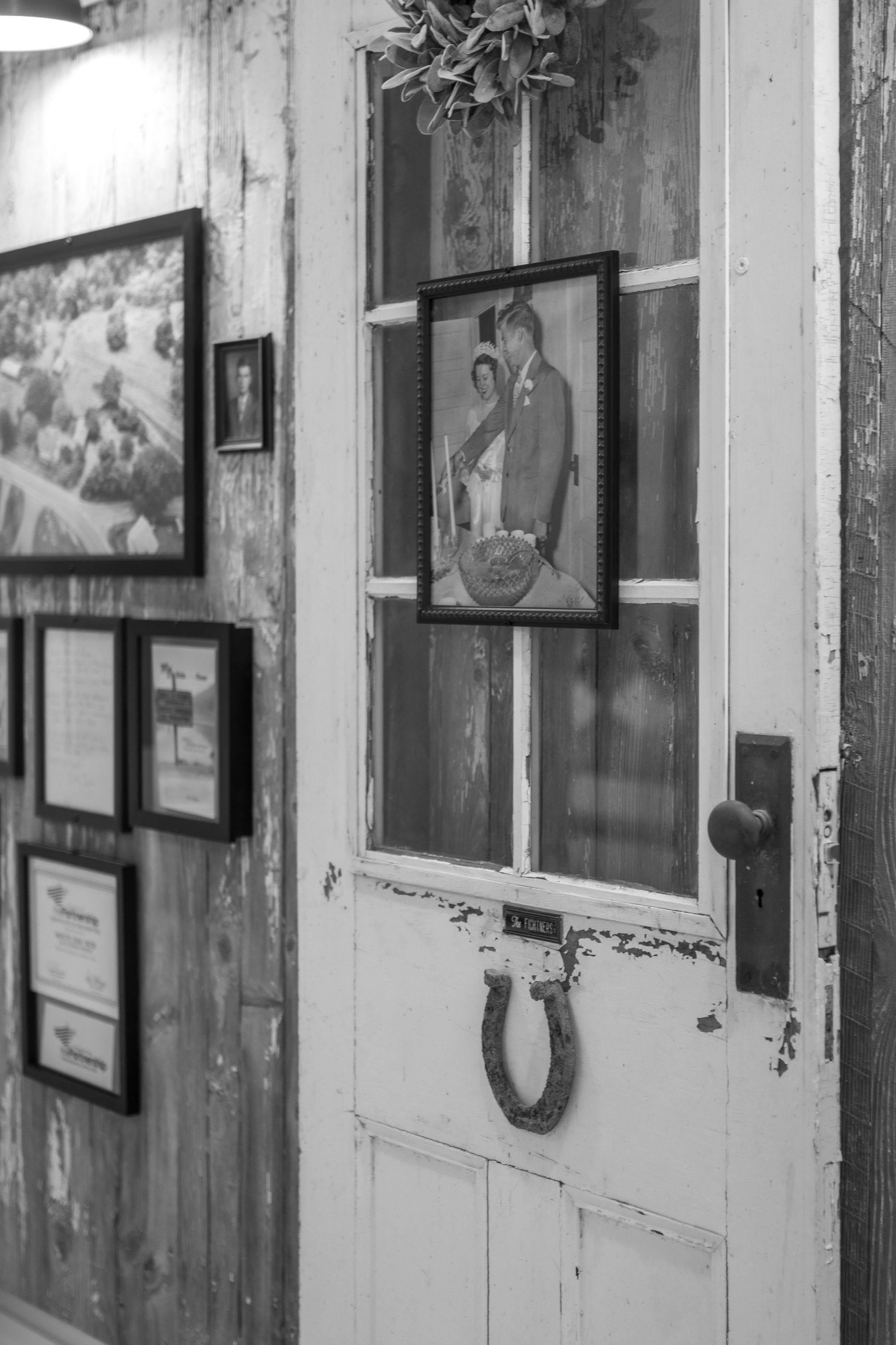 Weathered white door with framed portrait and wreath, beside a wall of framed pictures.