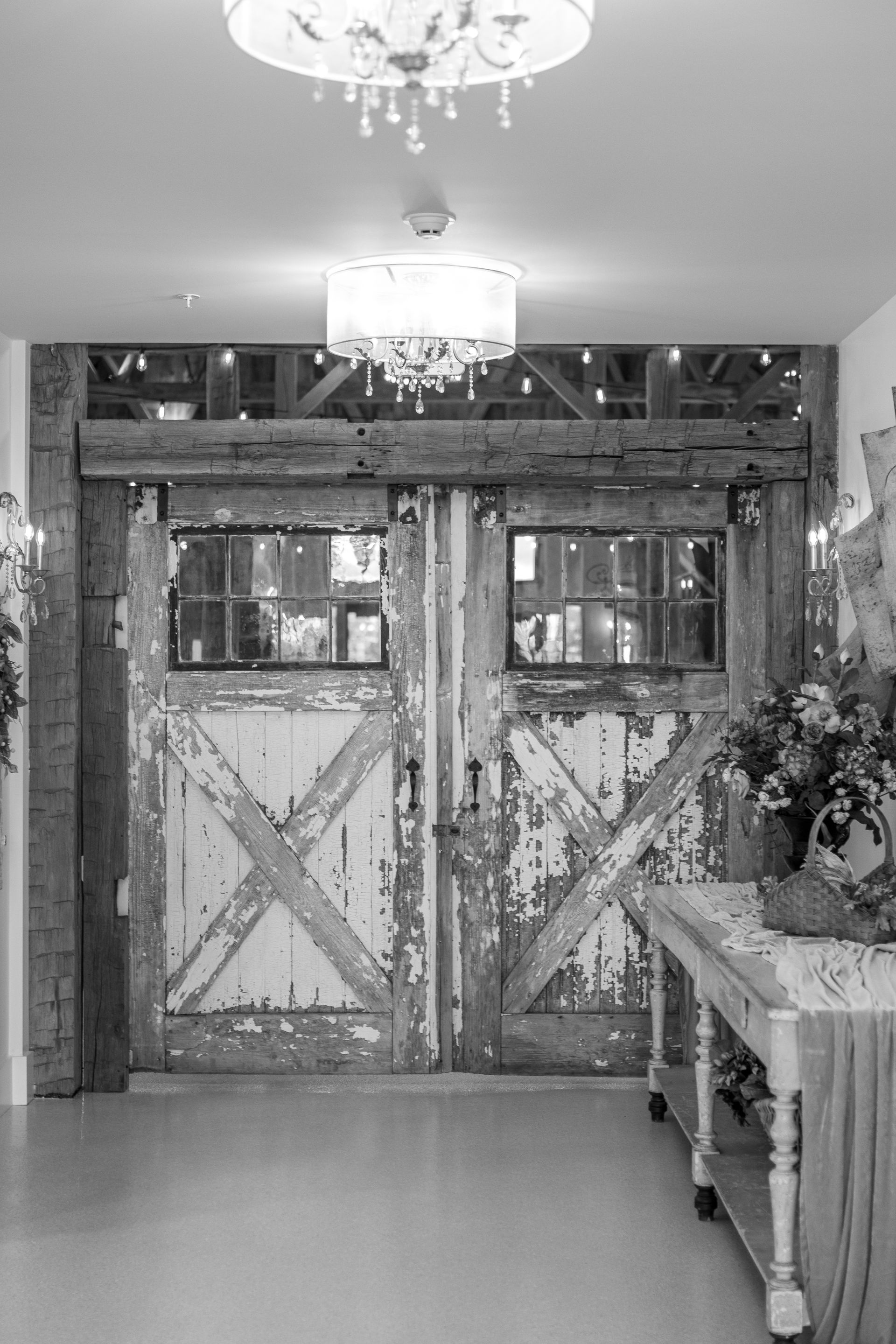 Rustic double barn doors with windows in a hallway, with chandeliers overhead and a table with flowers on the right.