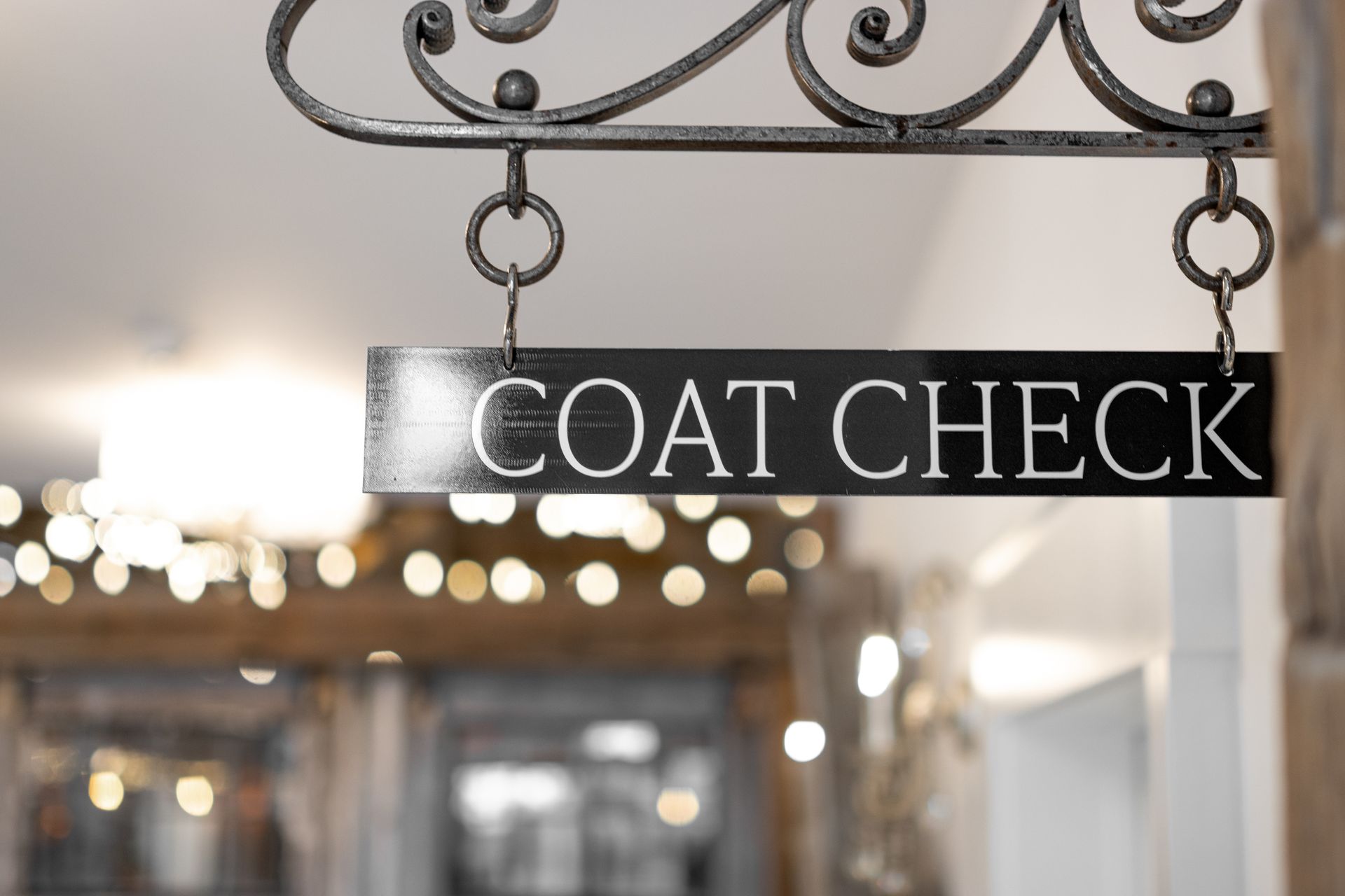 Black “COAT CHECK” sign hanging from an ornate metal frame in a softly lit indoor hallway
