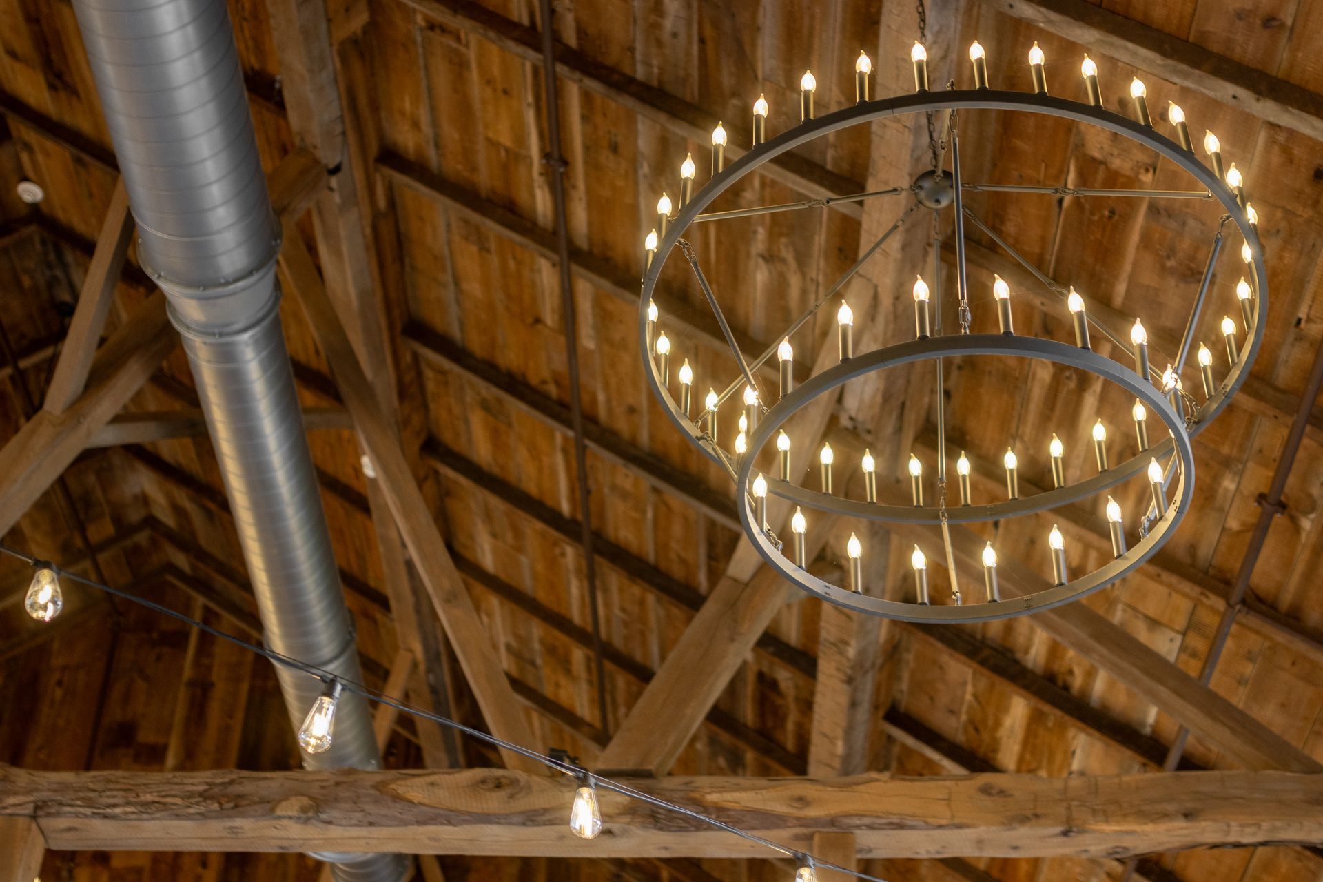 Rustic wooden ceiling with exposed beams, silver ductwork, and a large circular chandelier of hanging lights.