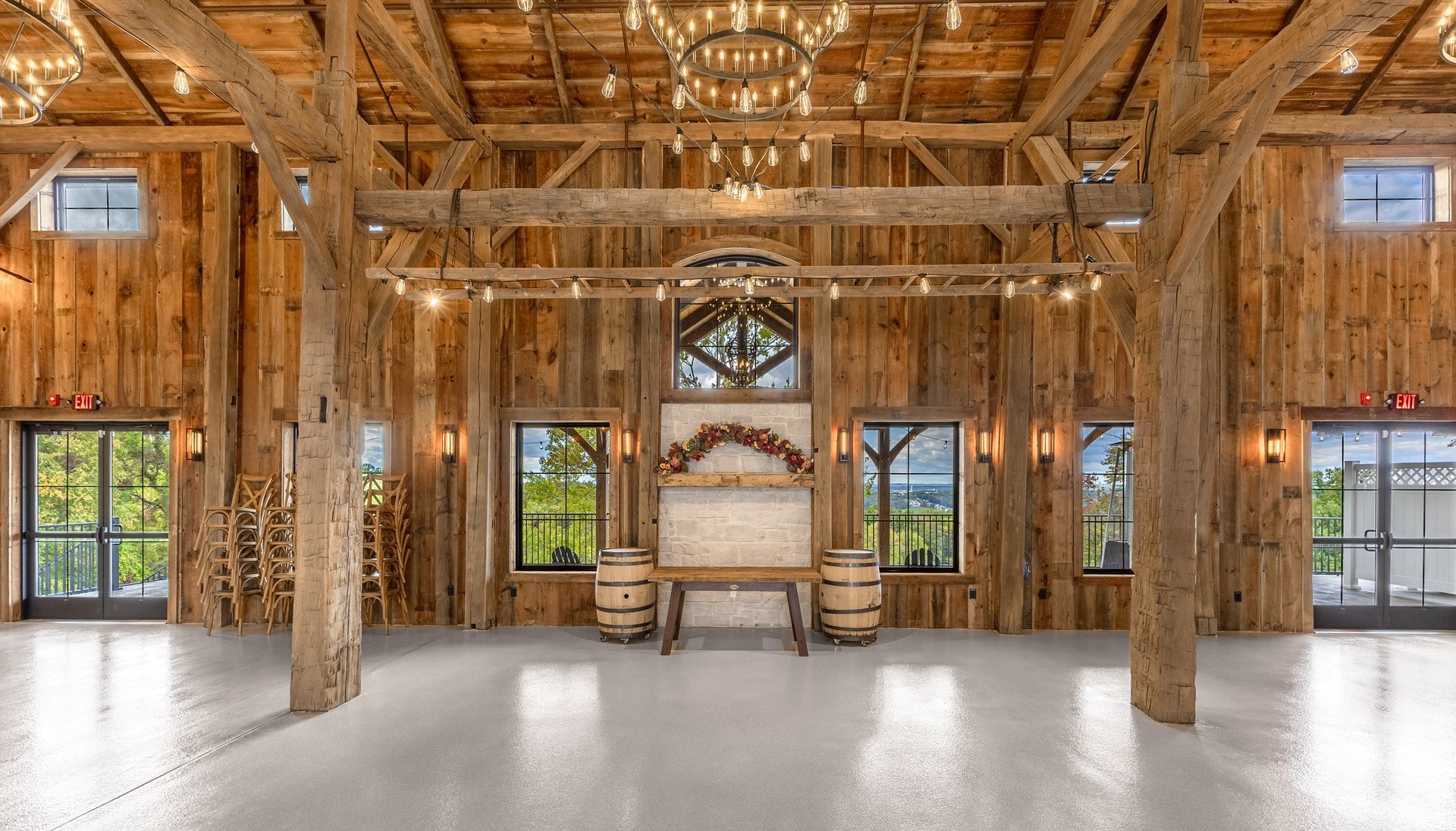 Rustic wooden hall with woven walls, large windows, hanging lights, and a central chandelier around a fire pit