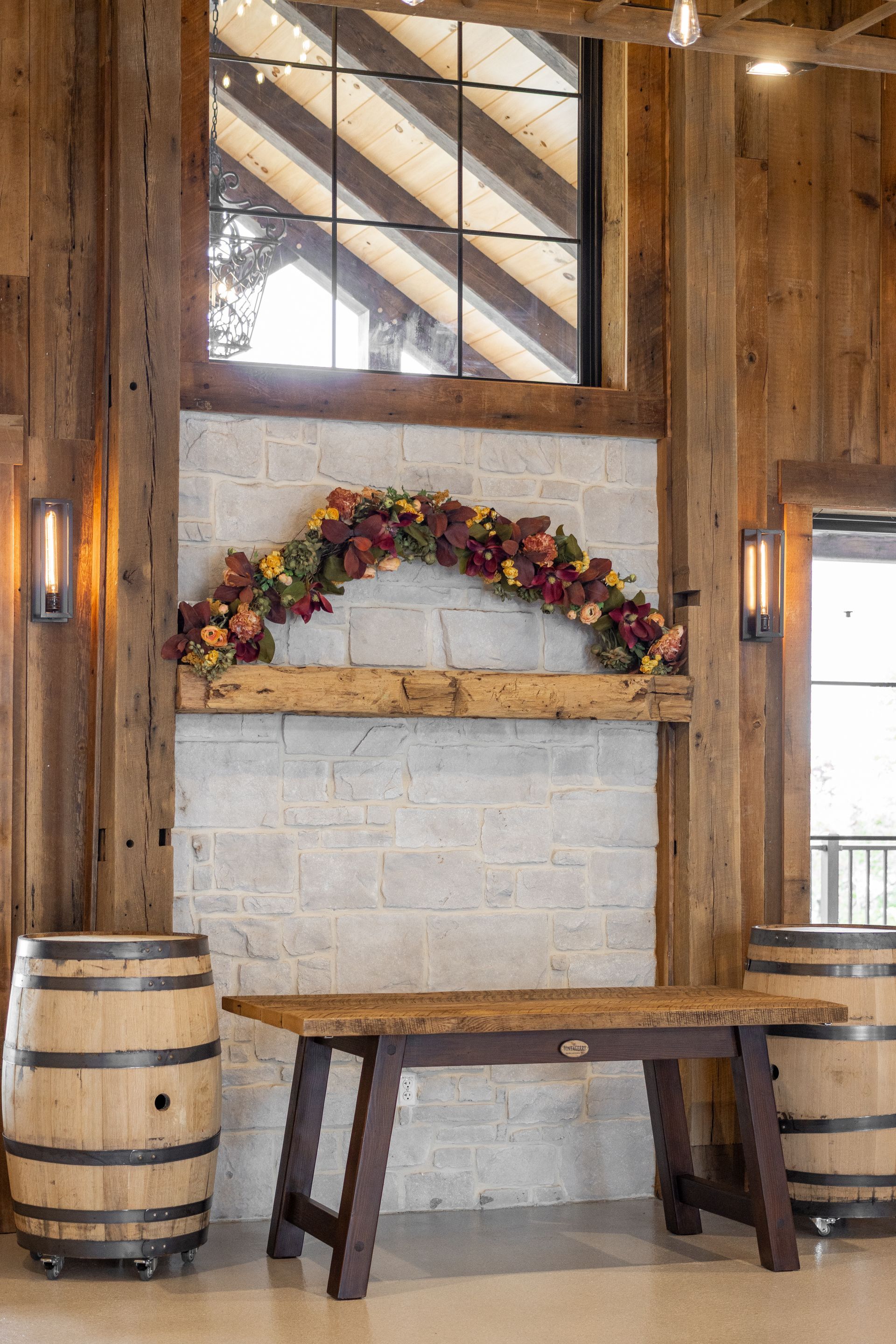 Rustic wood interior with a floral wreath above a mantel, bench, and two barrel tables