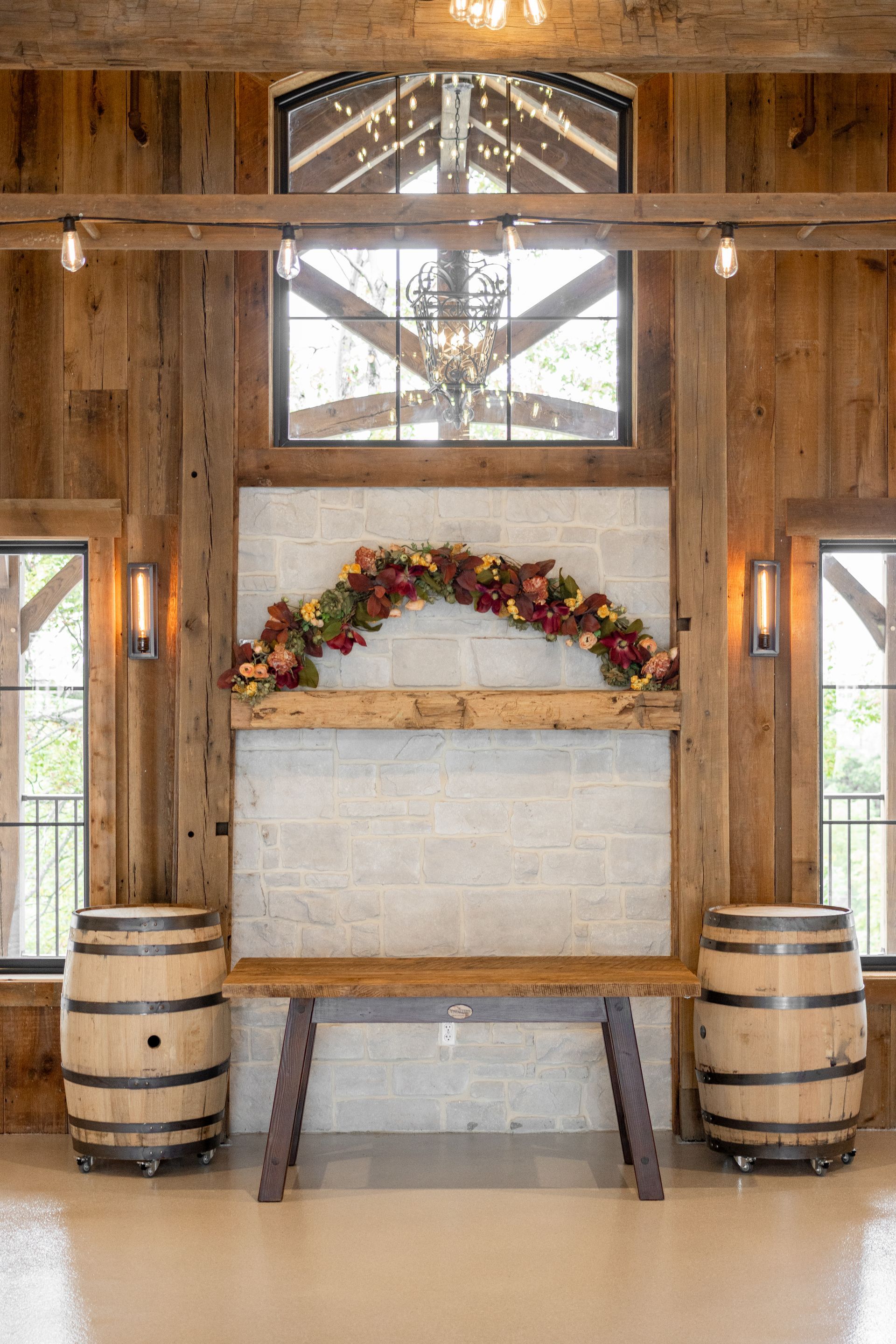 Rustic barn interior with a decorated mantel, barrels, and a bench beneath a window.