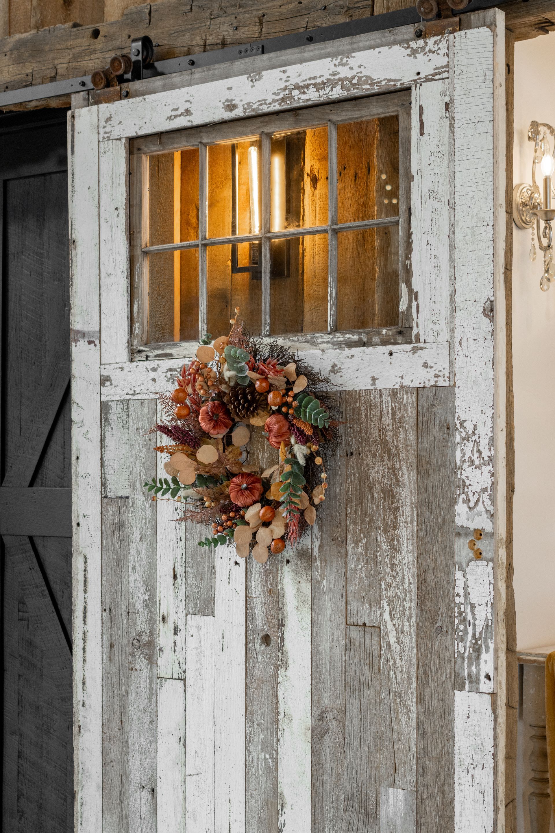 Weathered white door with a small window, warm interior light, and a floral wreath.