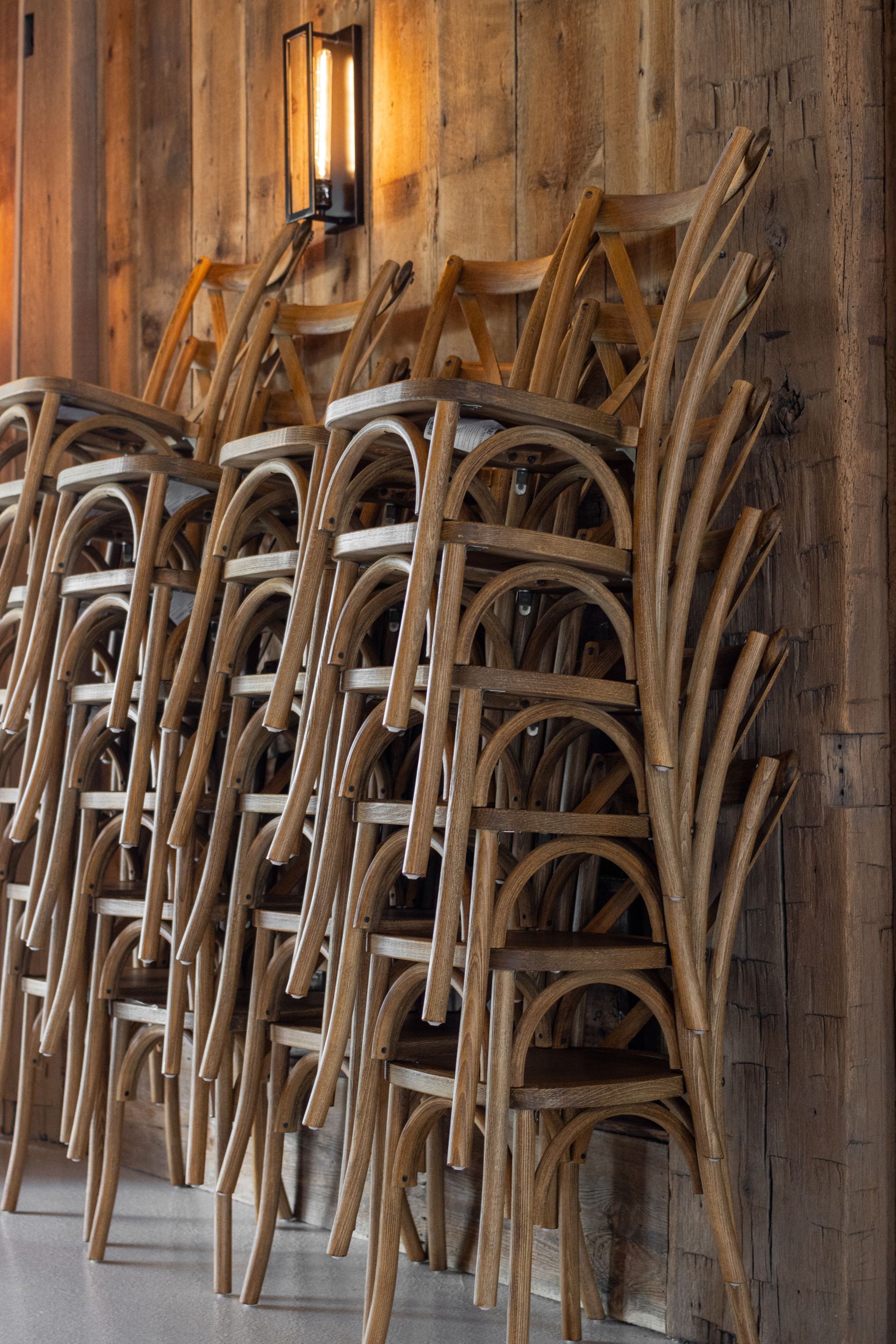 Stacked wooden chairs against a rustic wall with a warm wall sconce above them