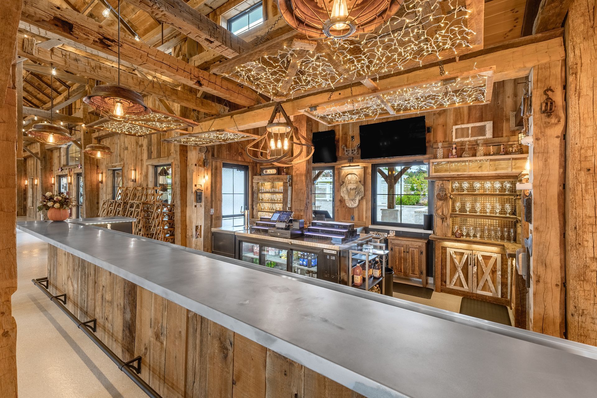 Rustic bar interior with a long stainless counter, warm string lights, wood decor, and a TV over the back bar