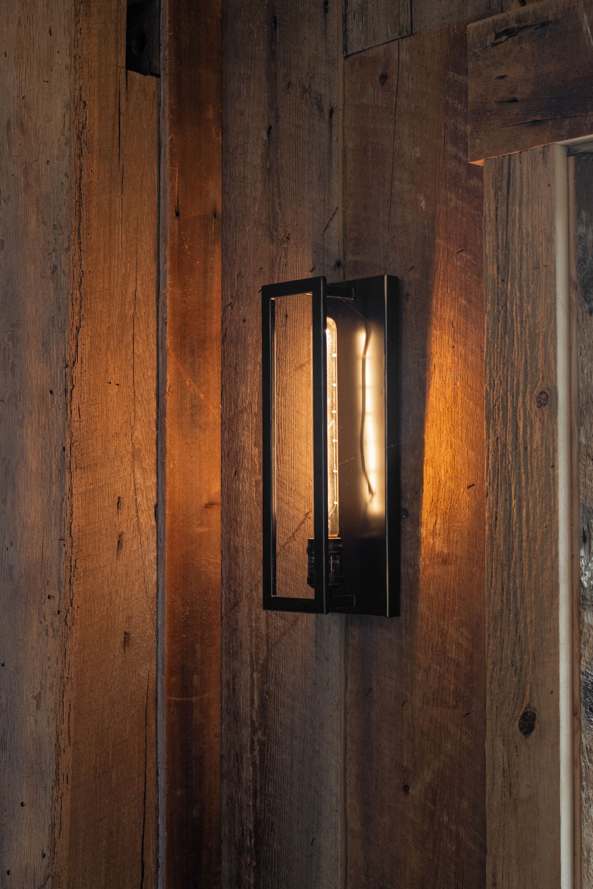 Black wall sconce glowing on a rustic wooden wall