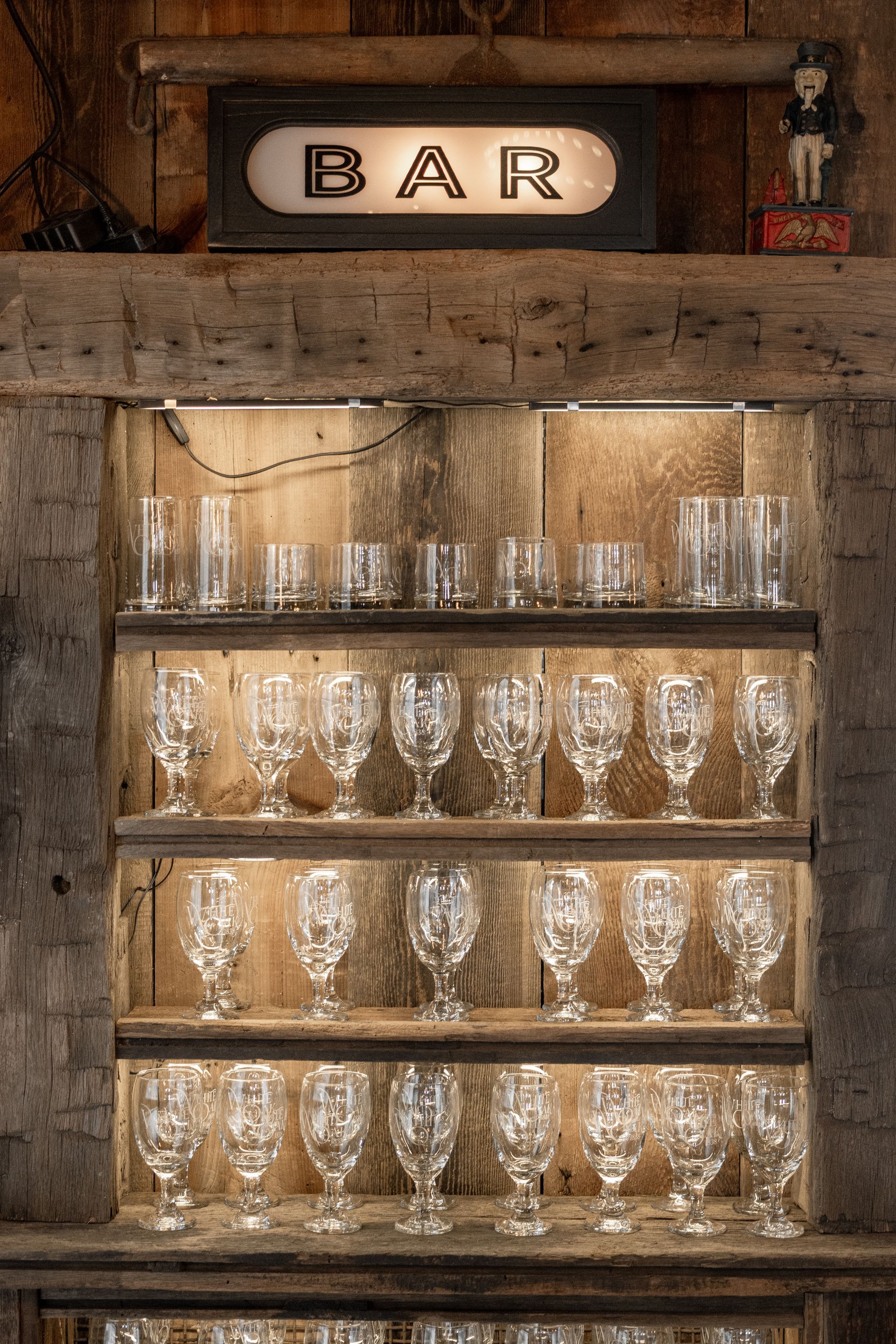 Rustic bar shelf with rows of empty glasses and a vintage “BAR” sign above.
