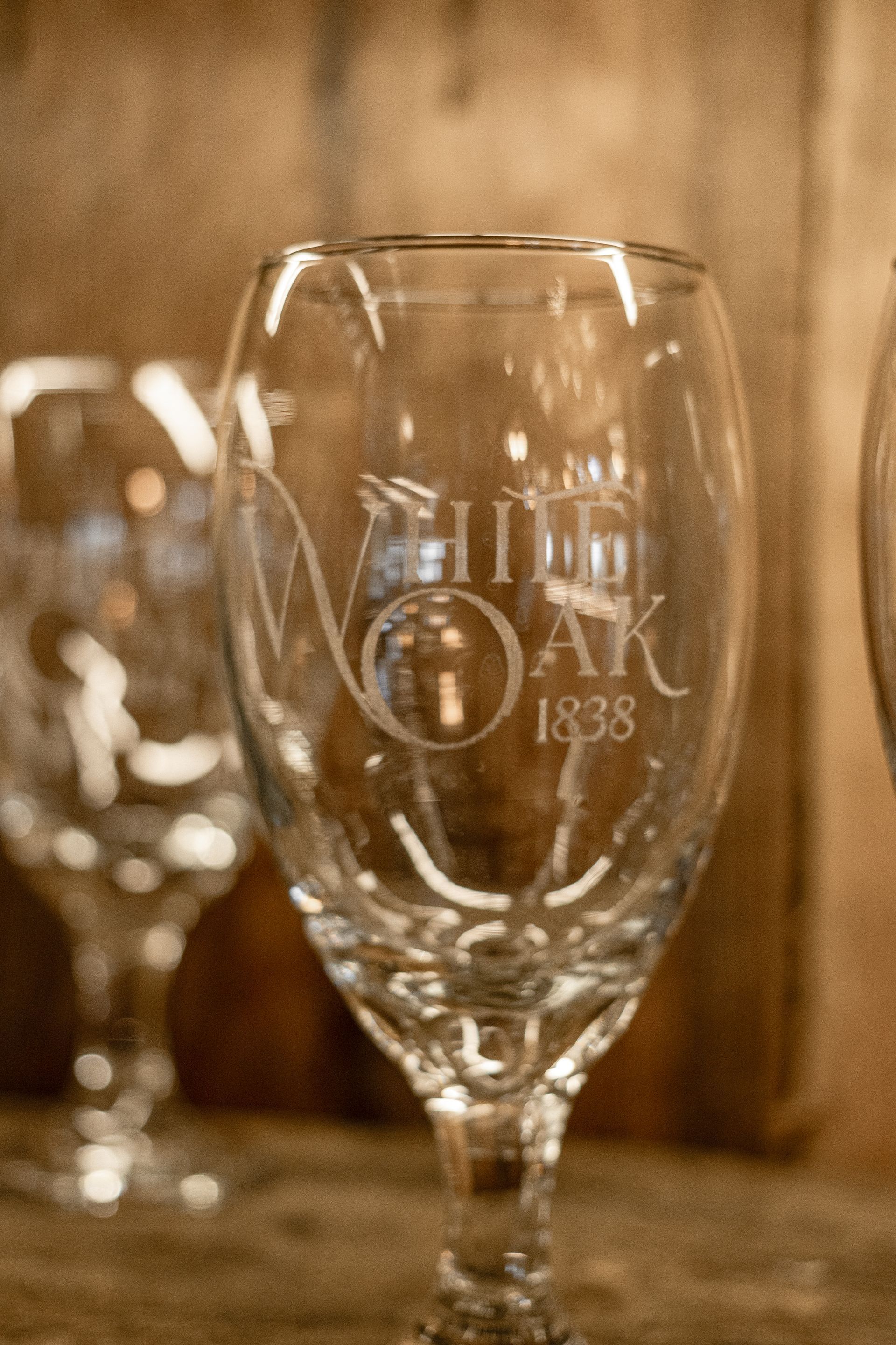 Close-up of a wine glass with “White Oak 1838” etched on it, against a warm wooden background.