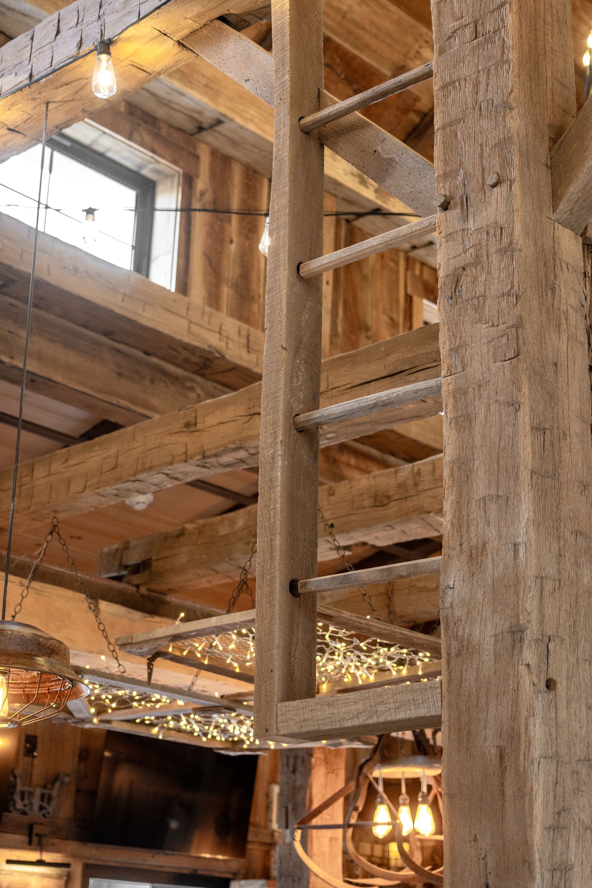 Rustic interior with exposed wooden beams, a ladder, and warm hanging lights in an unfinished loft.