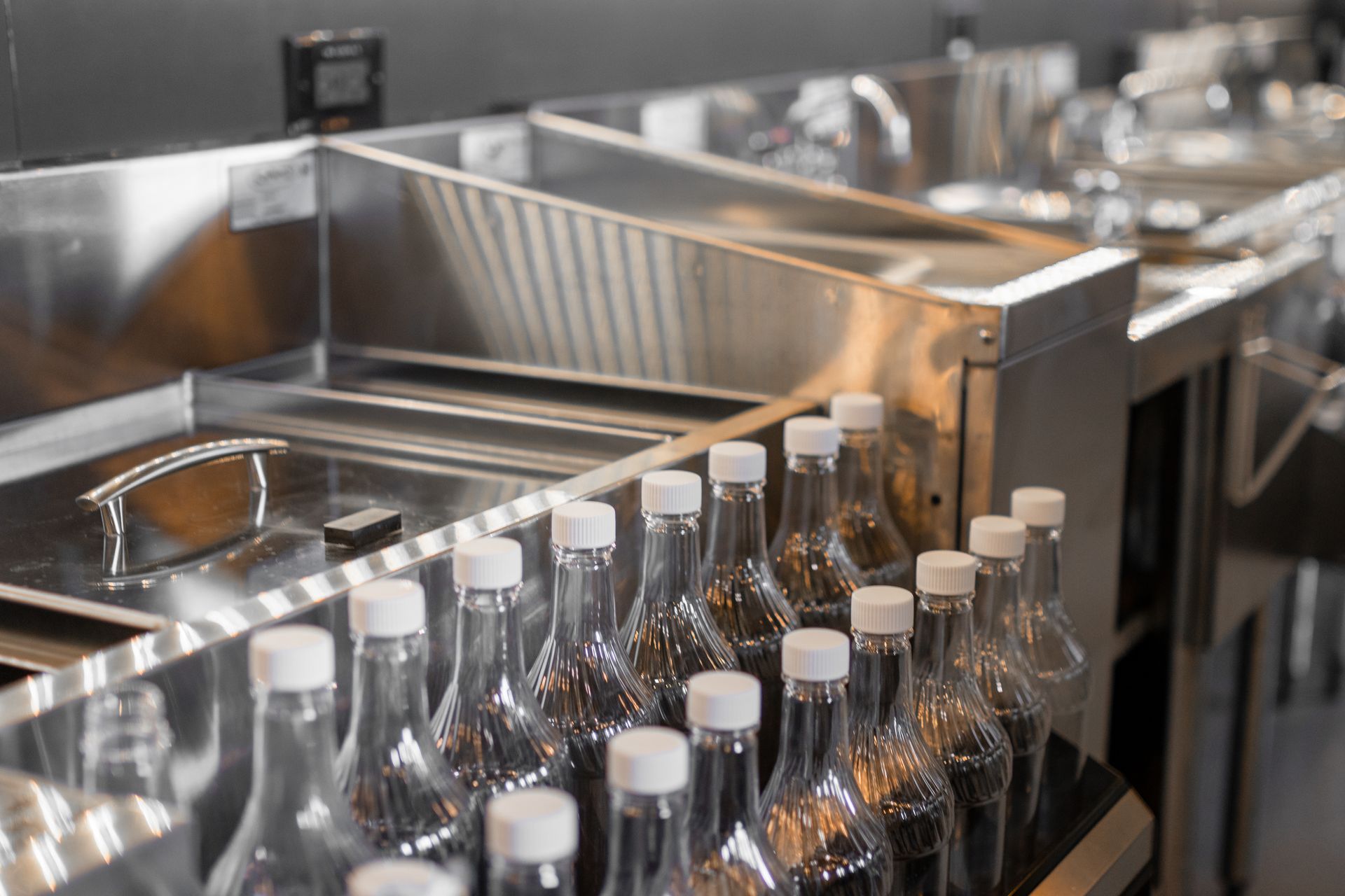 Rows of clear glass bottles in a stainless steel bottling line with metal trays and machinery