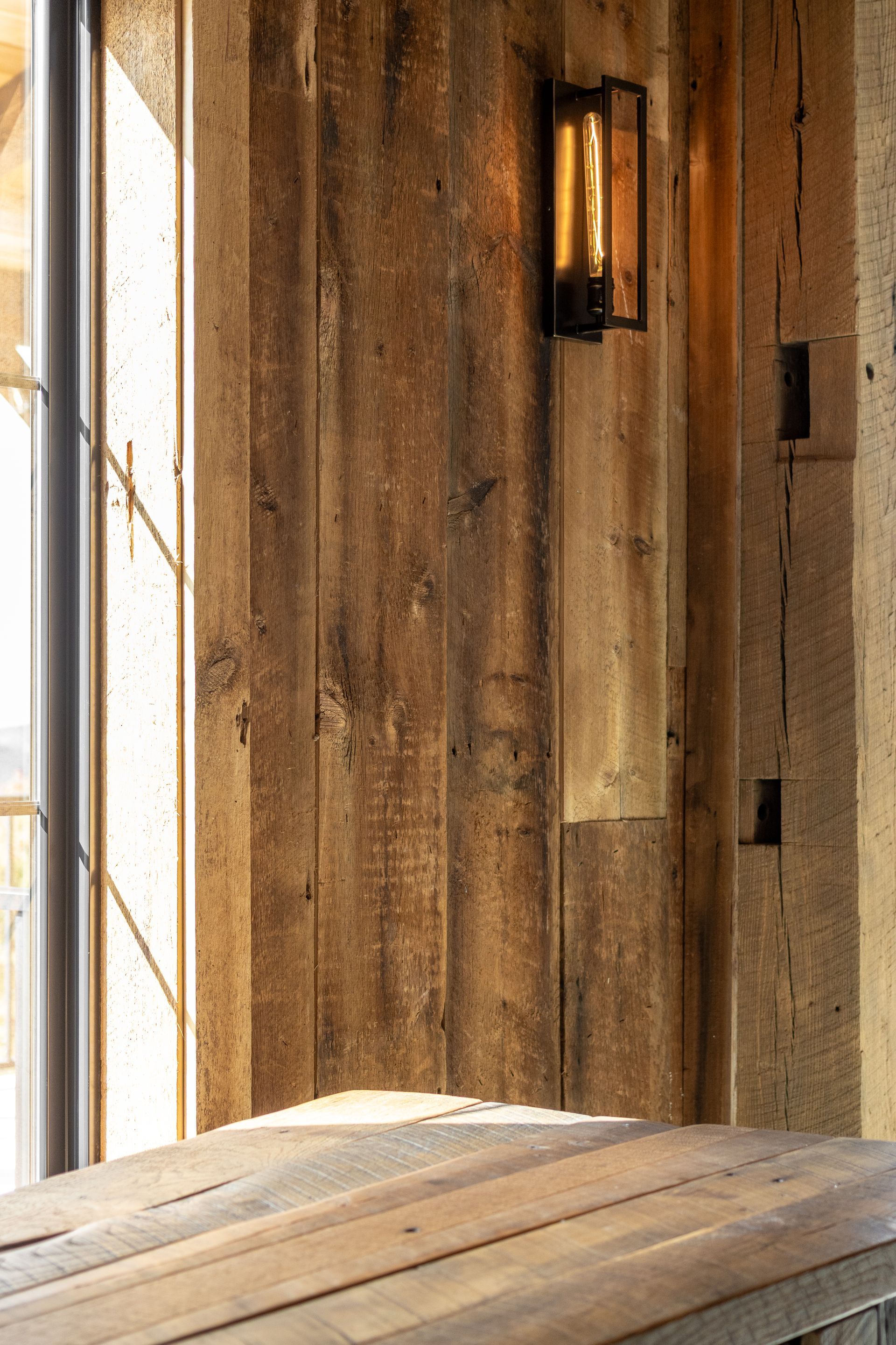 Rustic wooden wall with a lit wall sconce and sunlight on a wooden table by a window