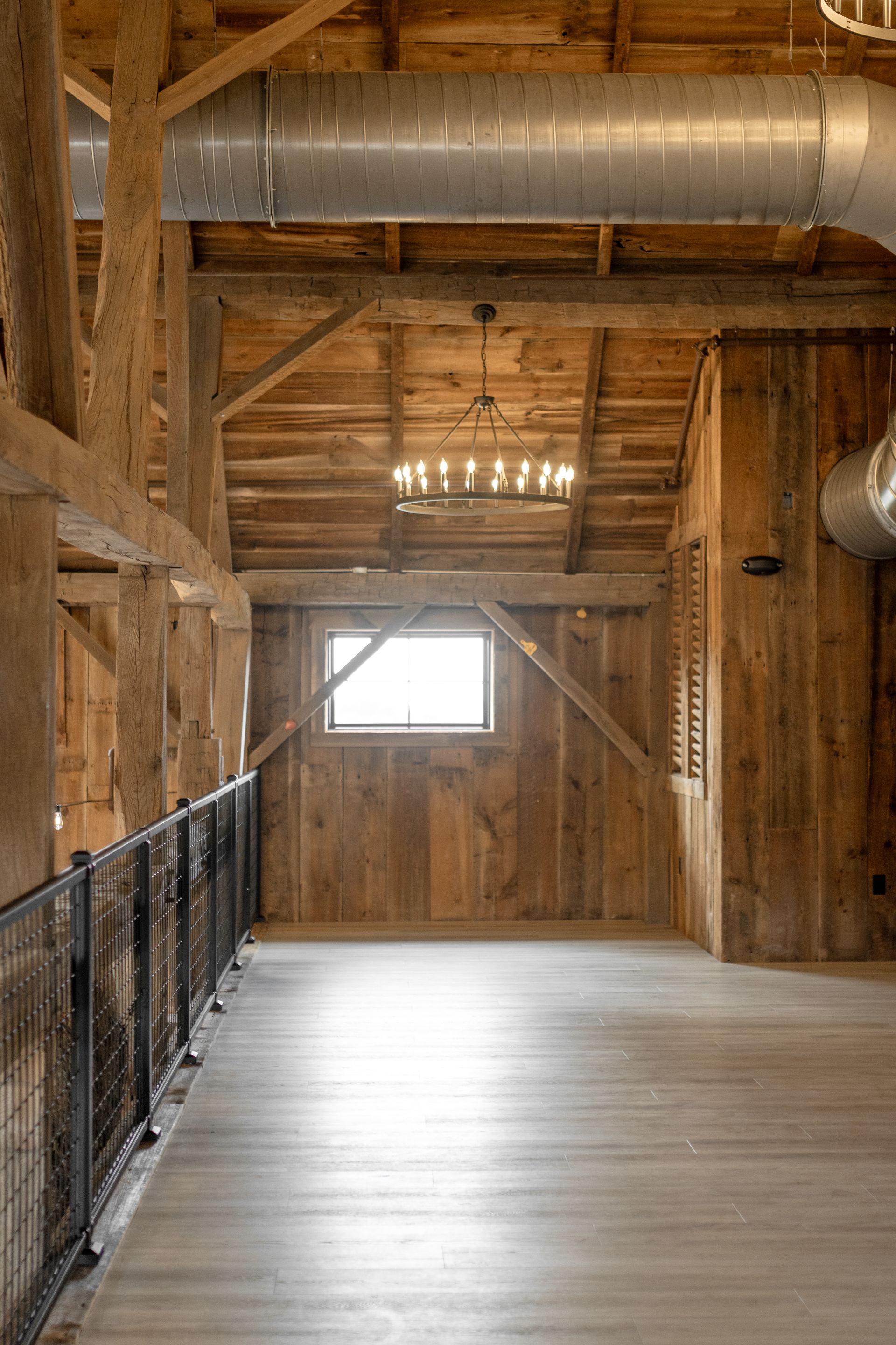 Rustic wooden loft hallway with metal railing, exposed beams, and a small window at the far end