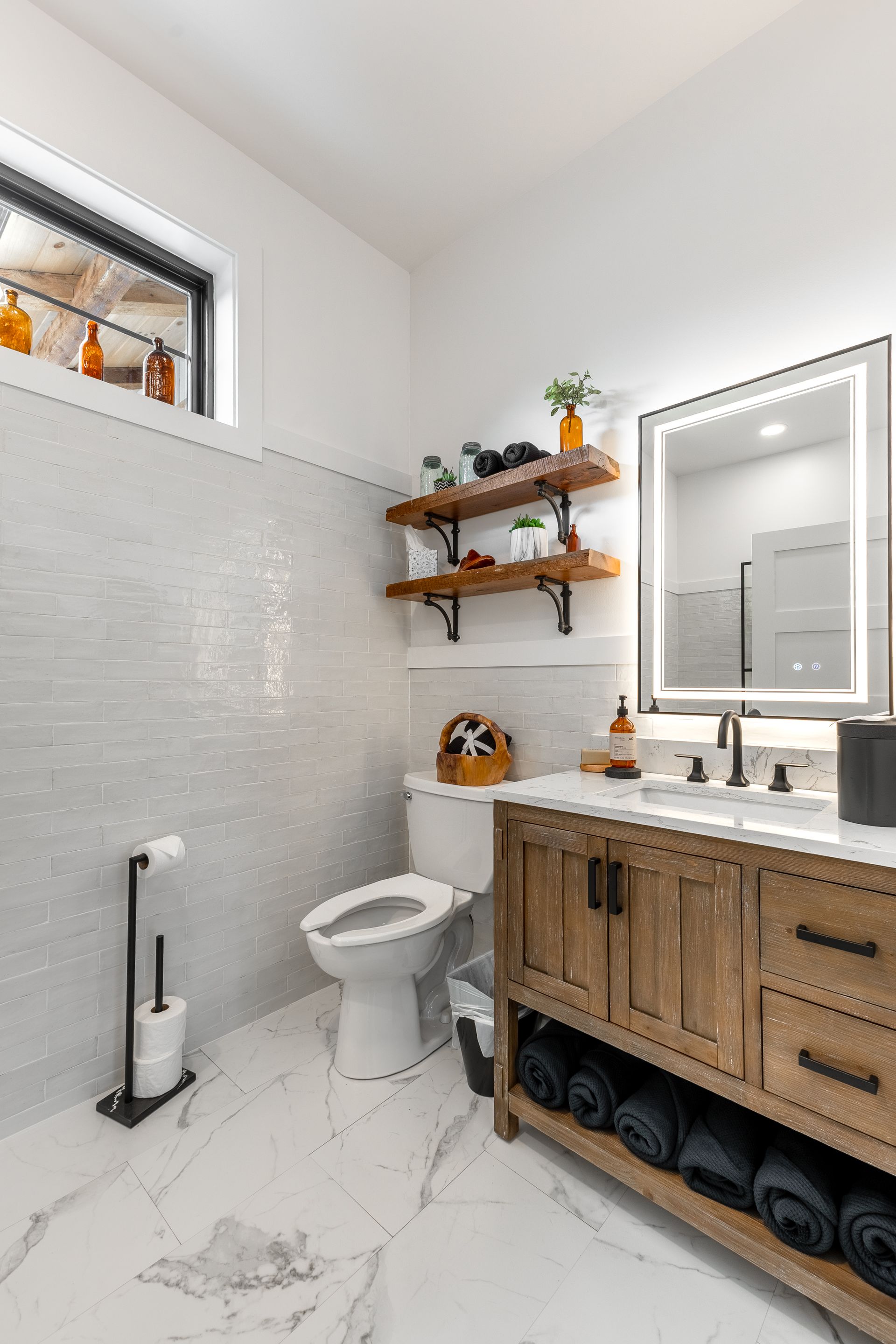 Modern bathroom with white walls, marble floor, wood vanity, toilet, mirror, and floating shelves
