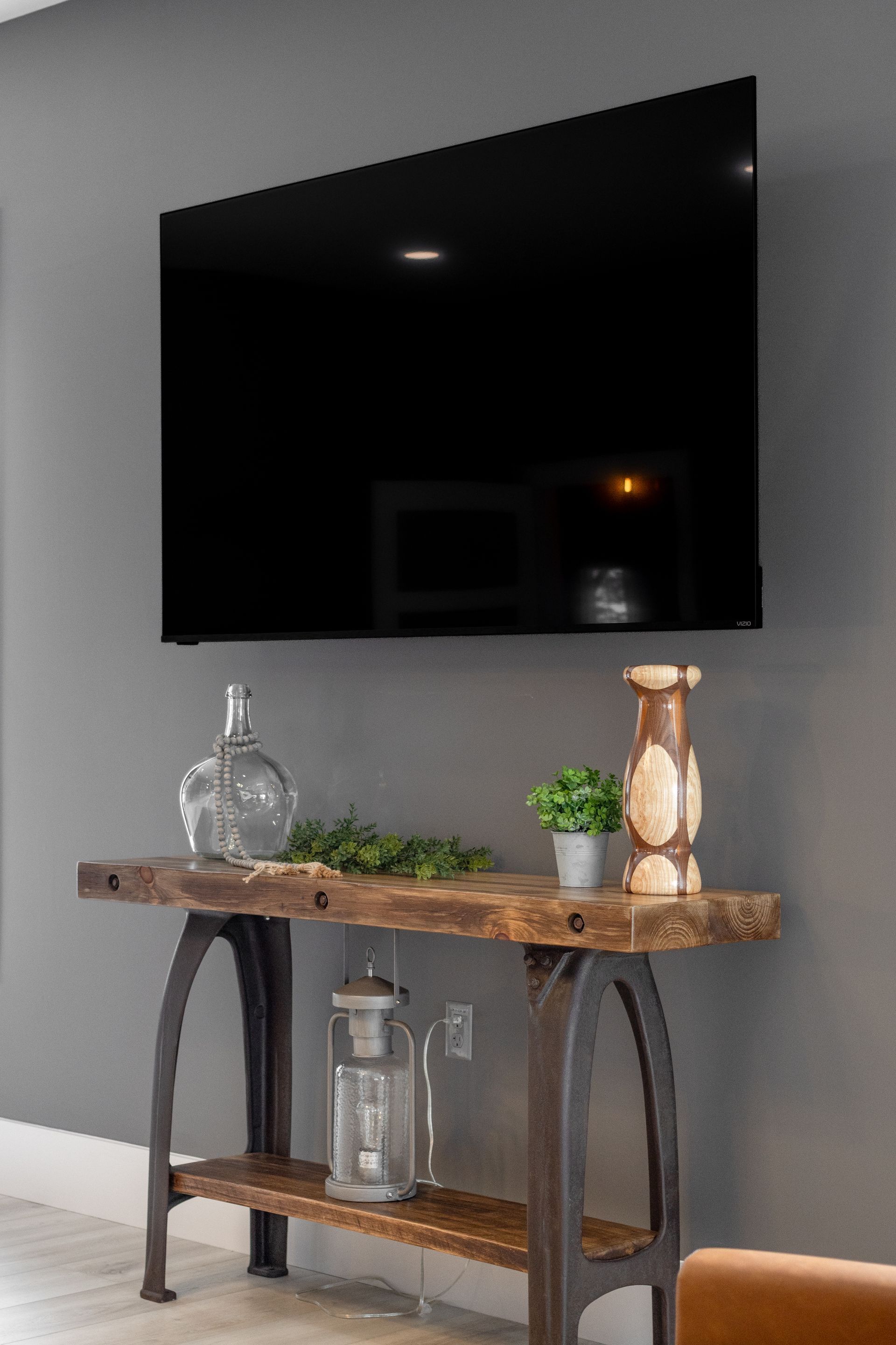 Wall-mounted TV above rustic wooden console table with glass decor and potted plants on a gray wall