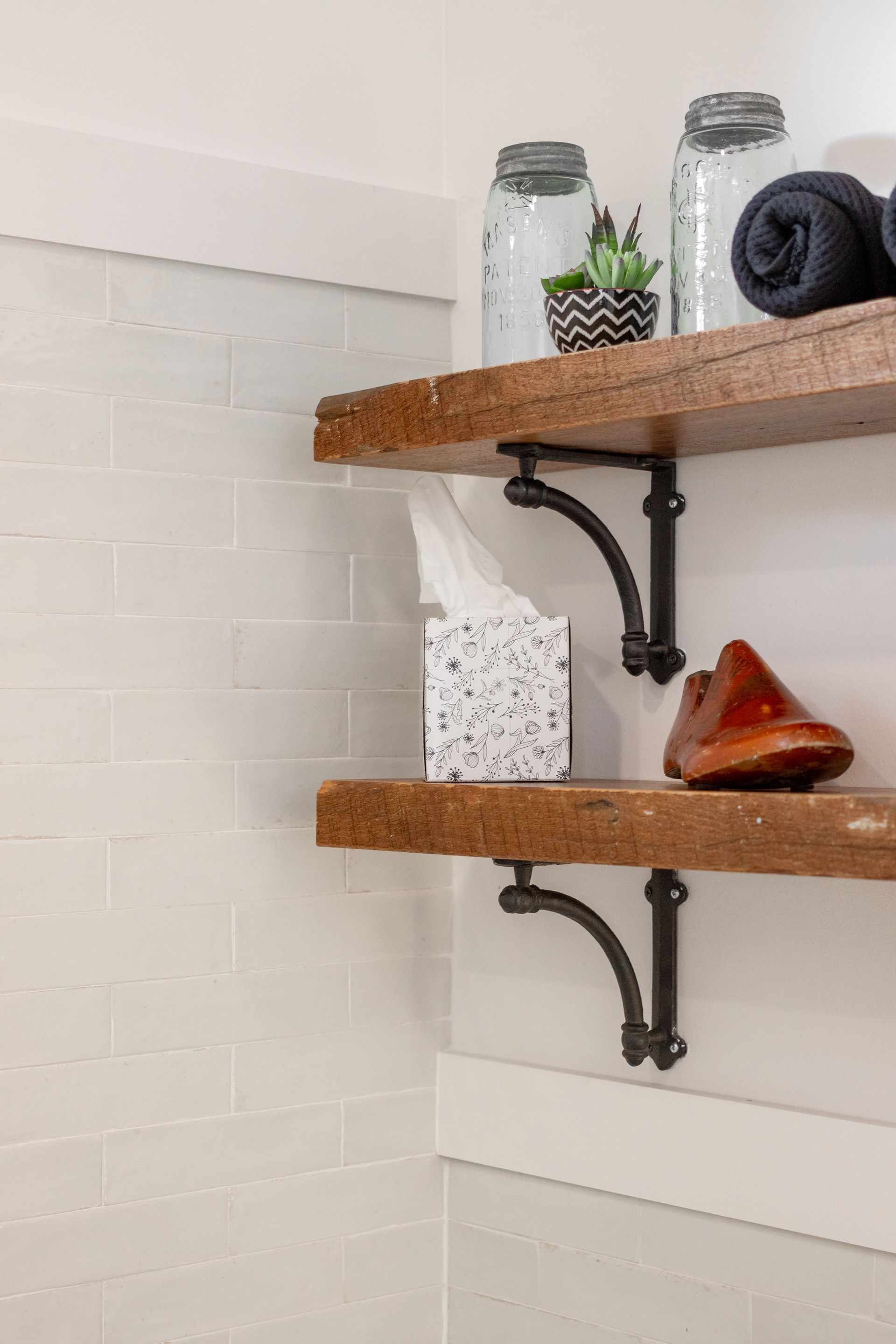 White bathroom wall with two wooden shelves holding jars, a folded towel, and toiletries.