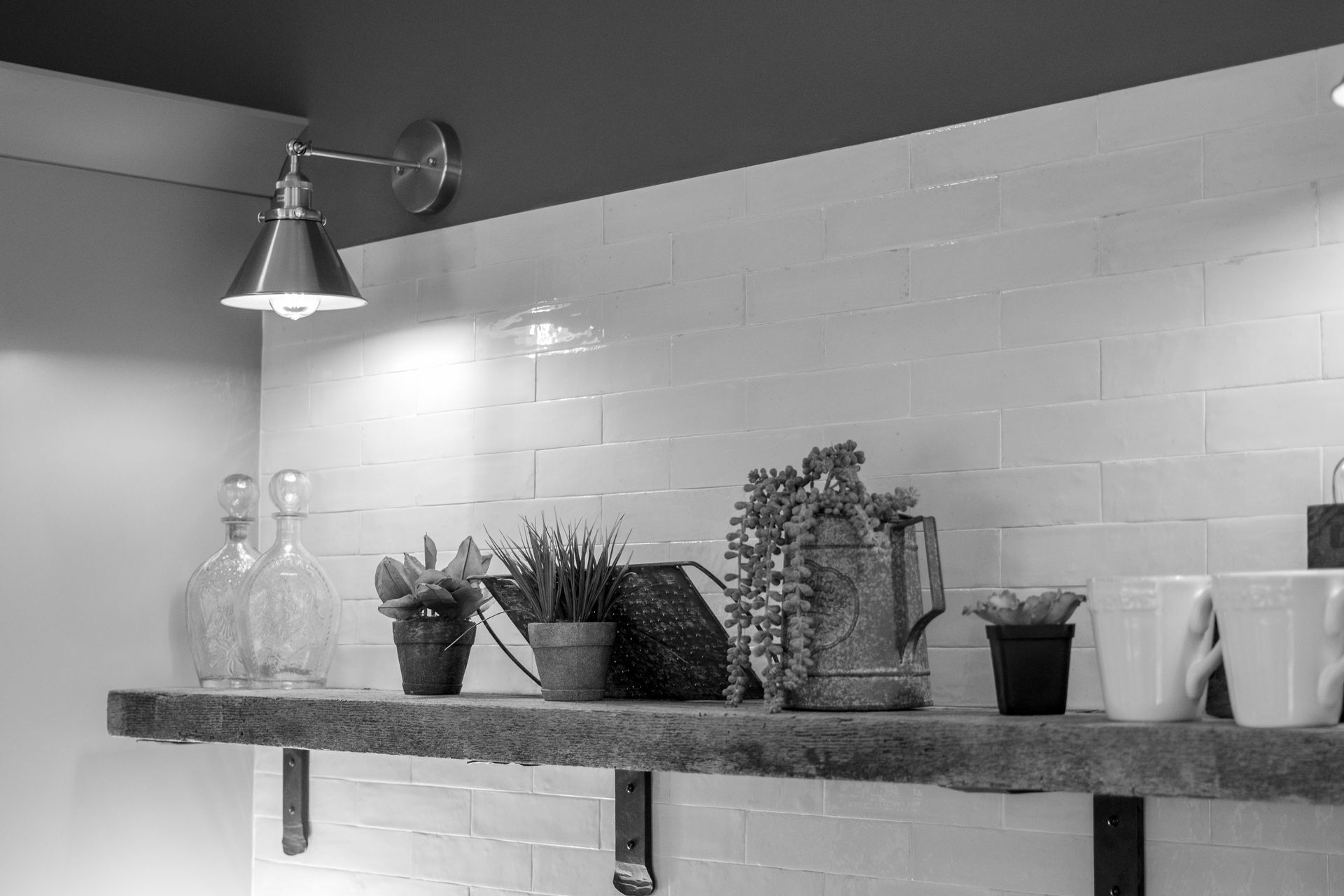 Wall shelf with potted plants and bottles under a hanging lamp against a tiled kitchen wall