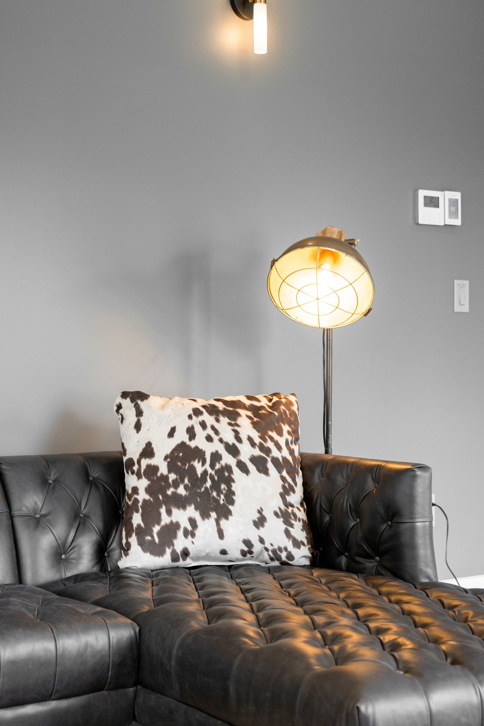 Modern living room with black leather sofa, patterned pillow, and lit floor lamp against gray wall