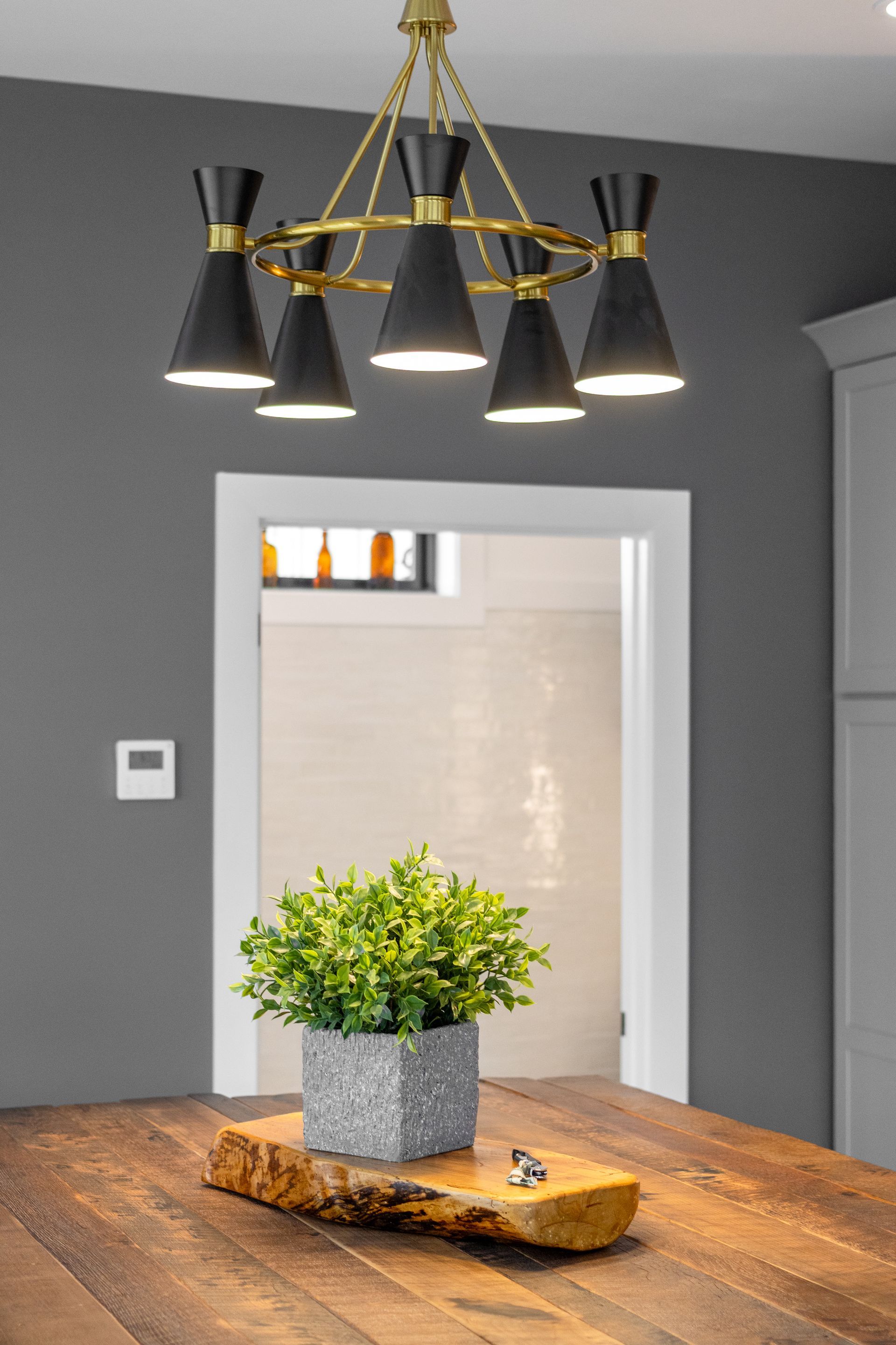 Modern dining room with a brass chandelier, wooden table, potted plant, and gray walls.