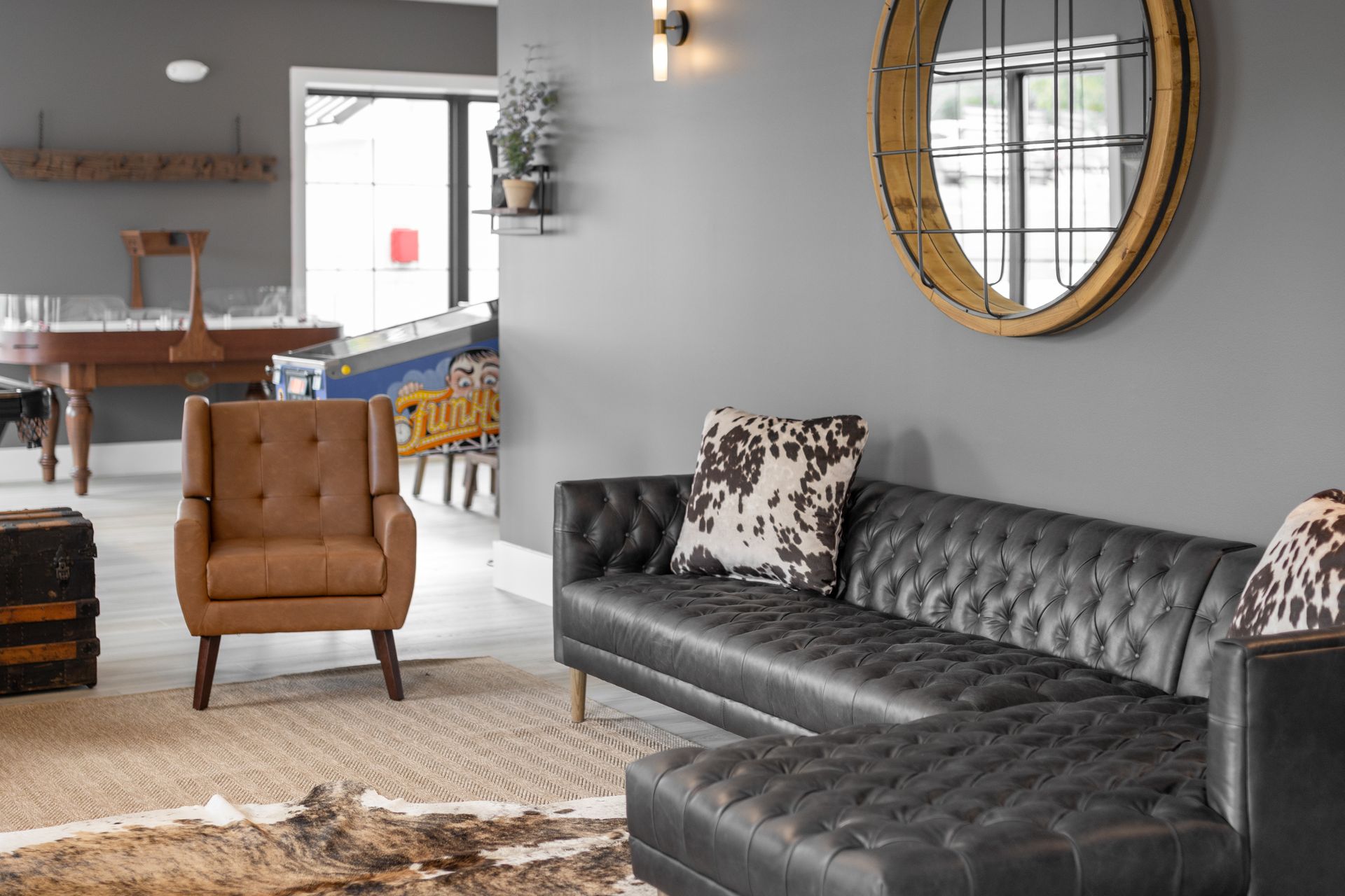 Modern living room with a black tufted sofa, tan chair, rug, and round mirror on a gray wall