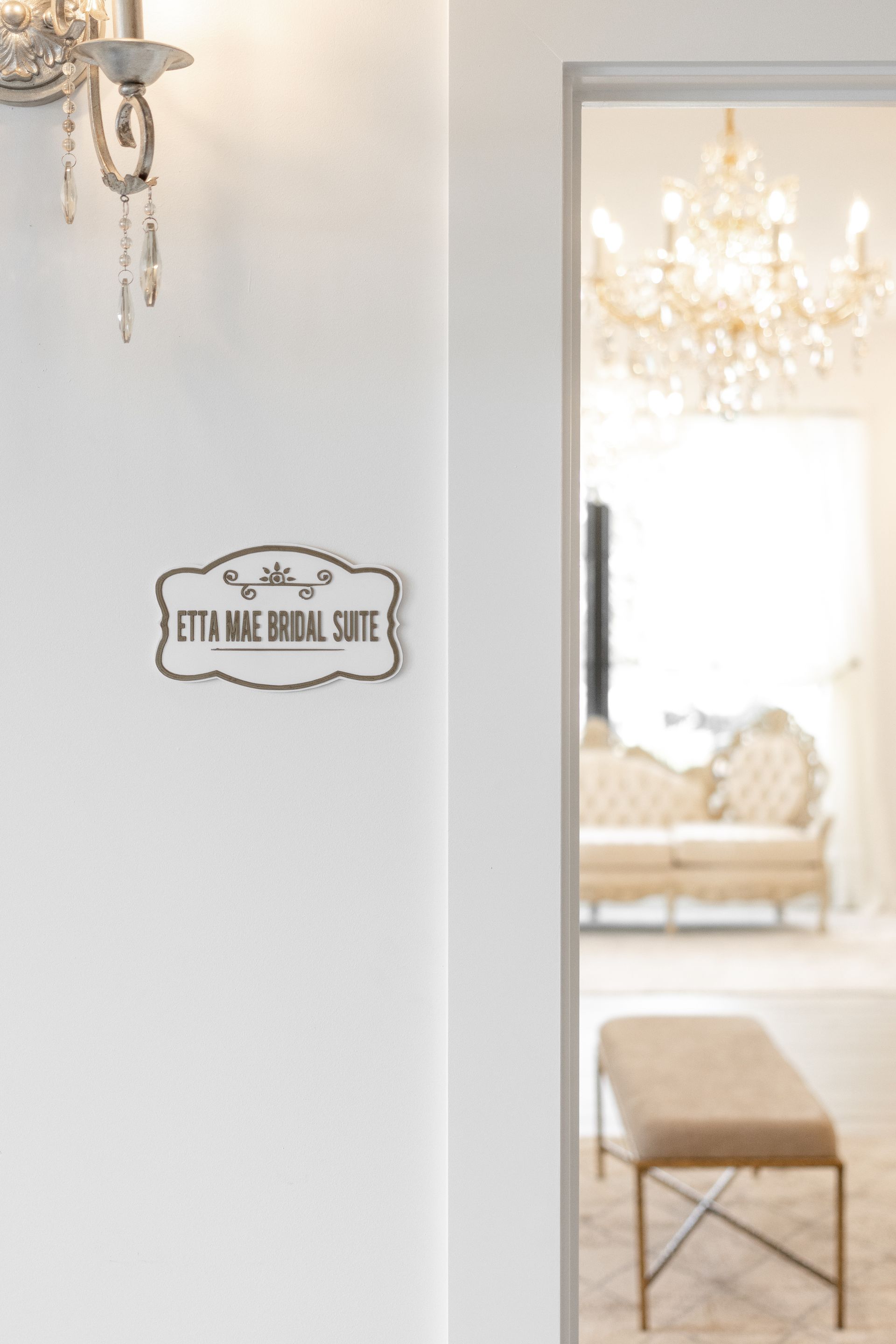 Elegant hallway with white walls, gold chandelier, and a bench visible through a doorway.