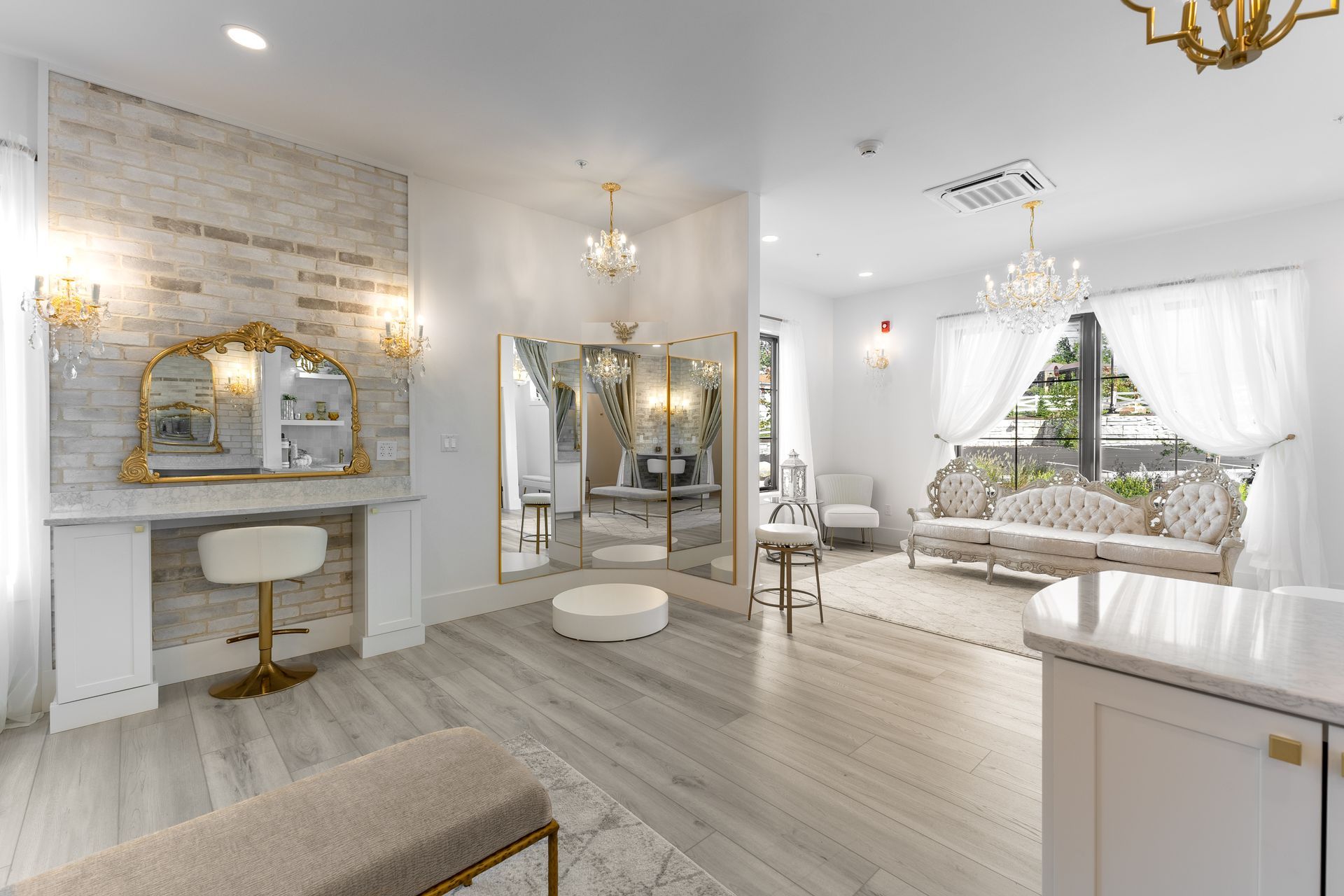 Elegant white vanity room with gold accents, chandelier lights, and a bright window seat.