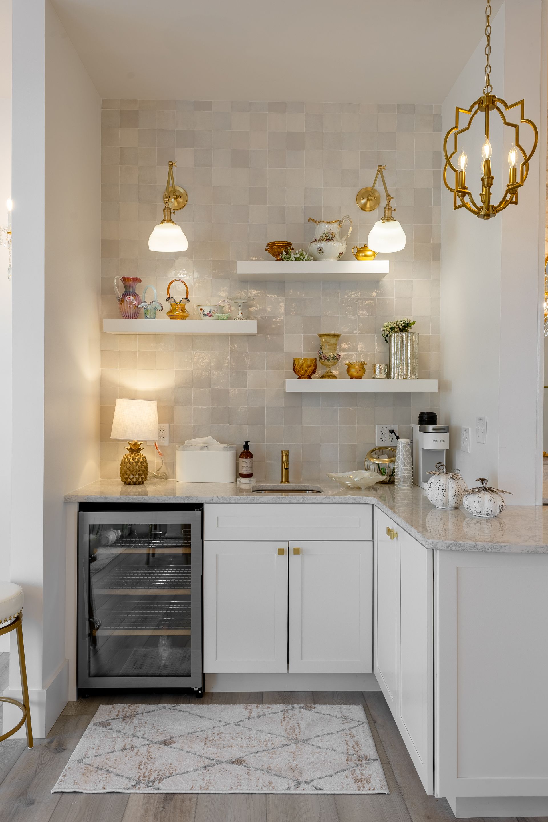 Bright, elegant kitchenette with white cabinets, floating shelves, gold pendant lights, and a small wine fridge.