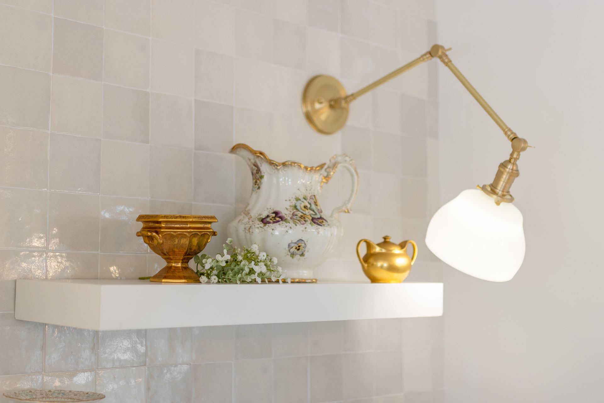 Decorative shelf with gold-toned glassware, a white pitcher, and a brass wall lamp against tiled wall