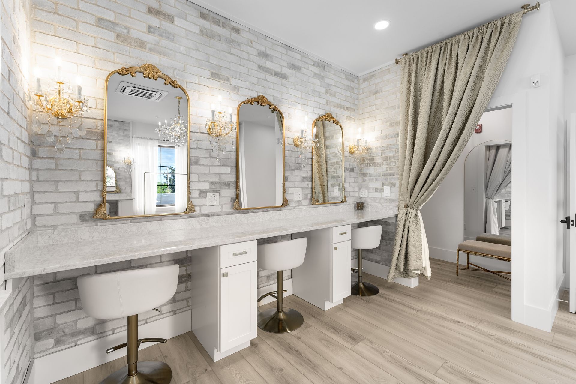 Bright salon station with gold-framed mirrors, white brick walls, stools, and floor-length curtains