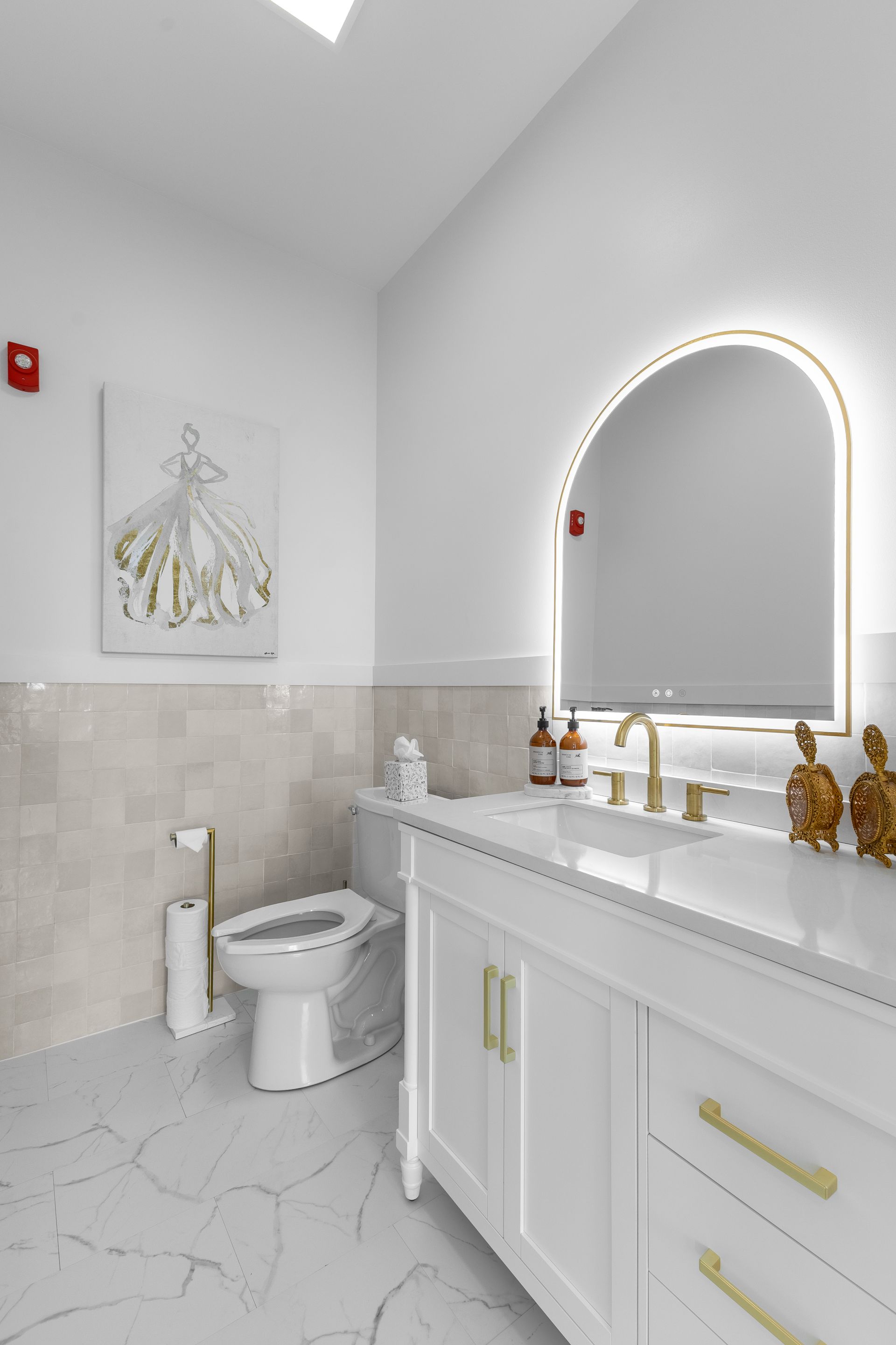 Bright white bathroom with marble vanity, arched lit mirror, gold accents, and a toilet on the left