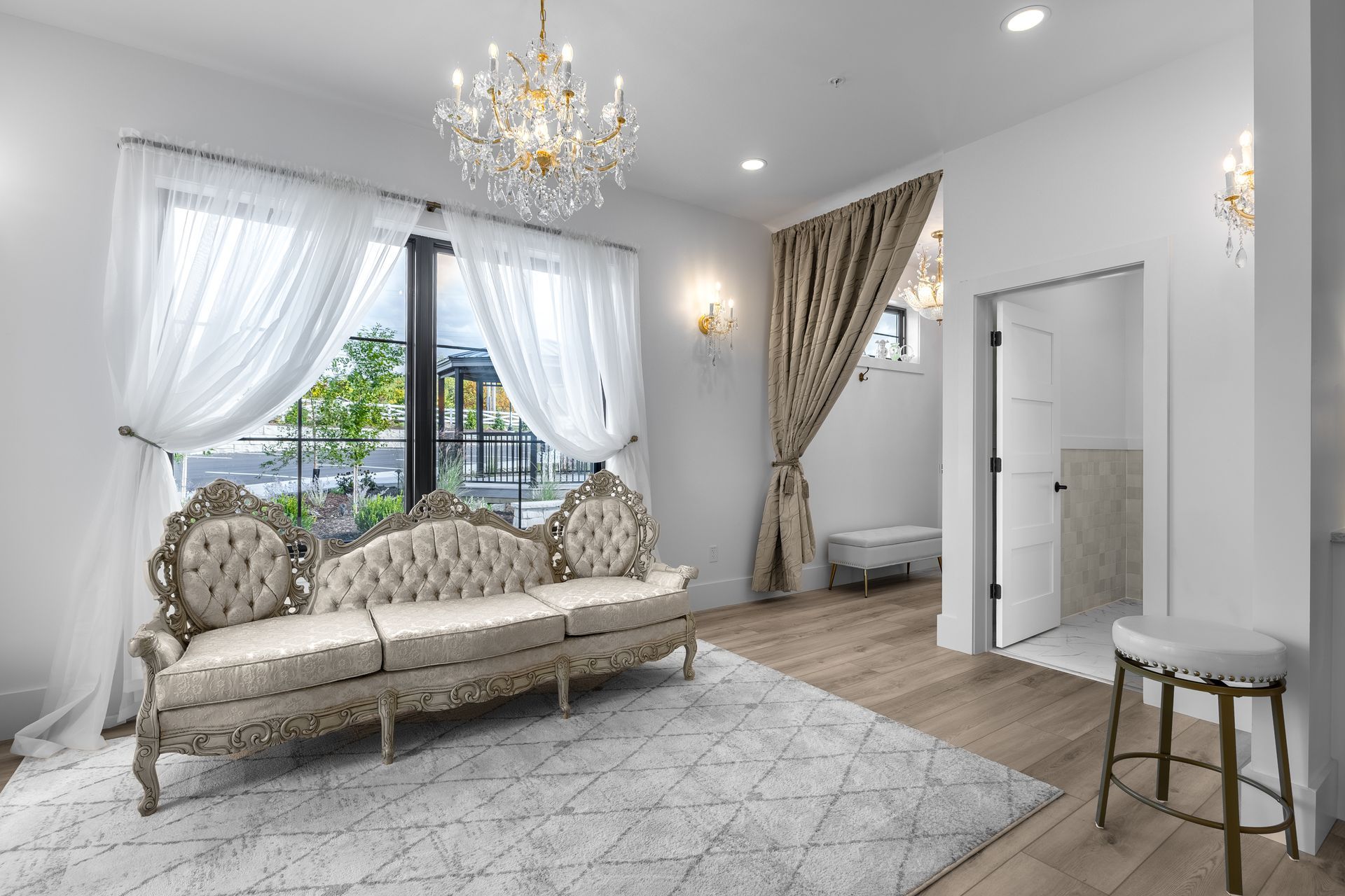 Elegant white living room with ornate sofa, chandelier, sheer curtains, and large window overlooking greenery