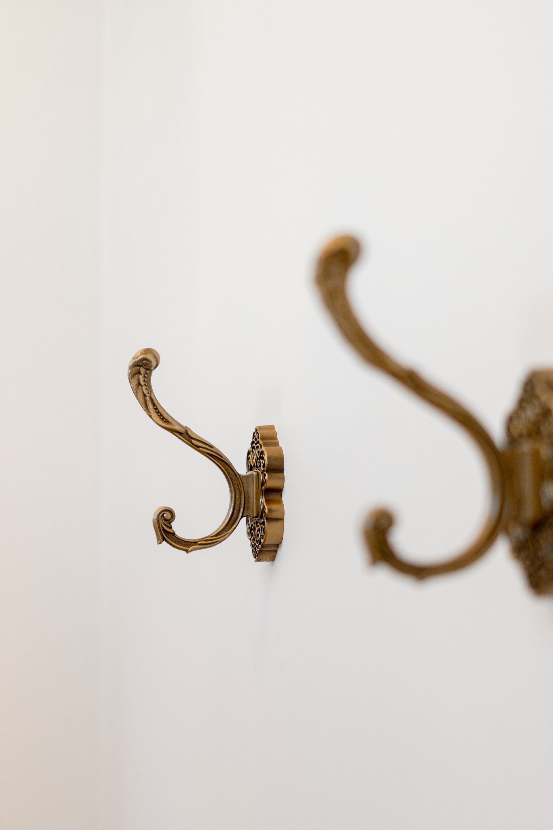 Close-up of ornate brass wall hooks mounted on a white wall, with one hook in focus and another blurred in the background