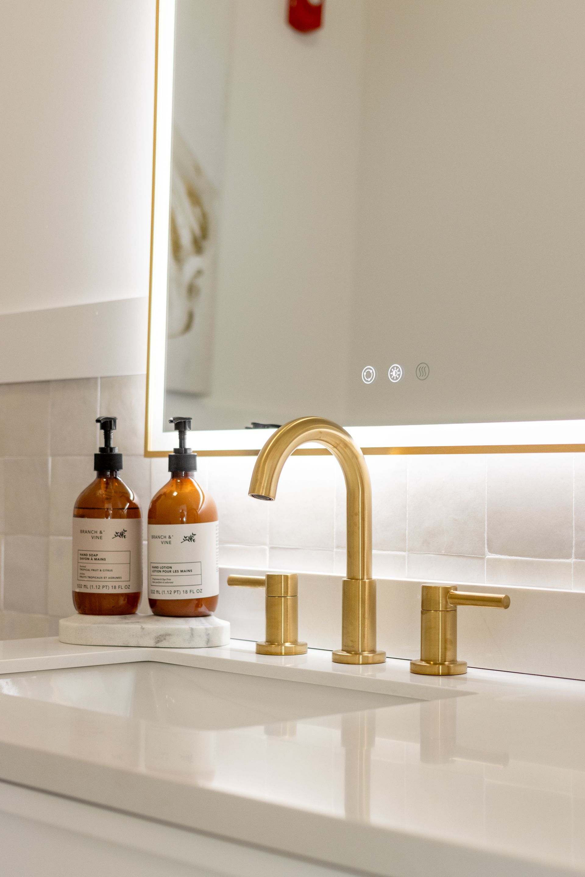 Gold faucet and soap dispensers on a white bathroom vanity beside a mirror and lighted backsplash