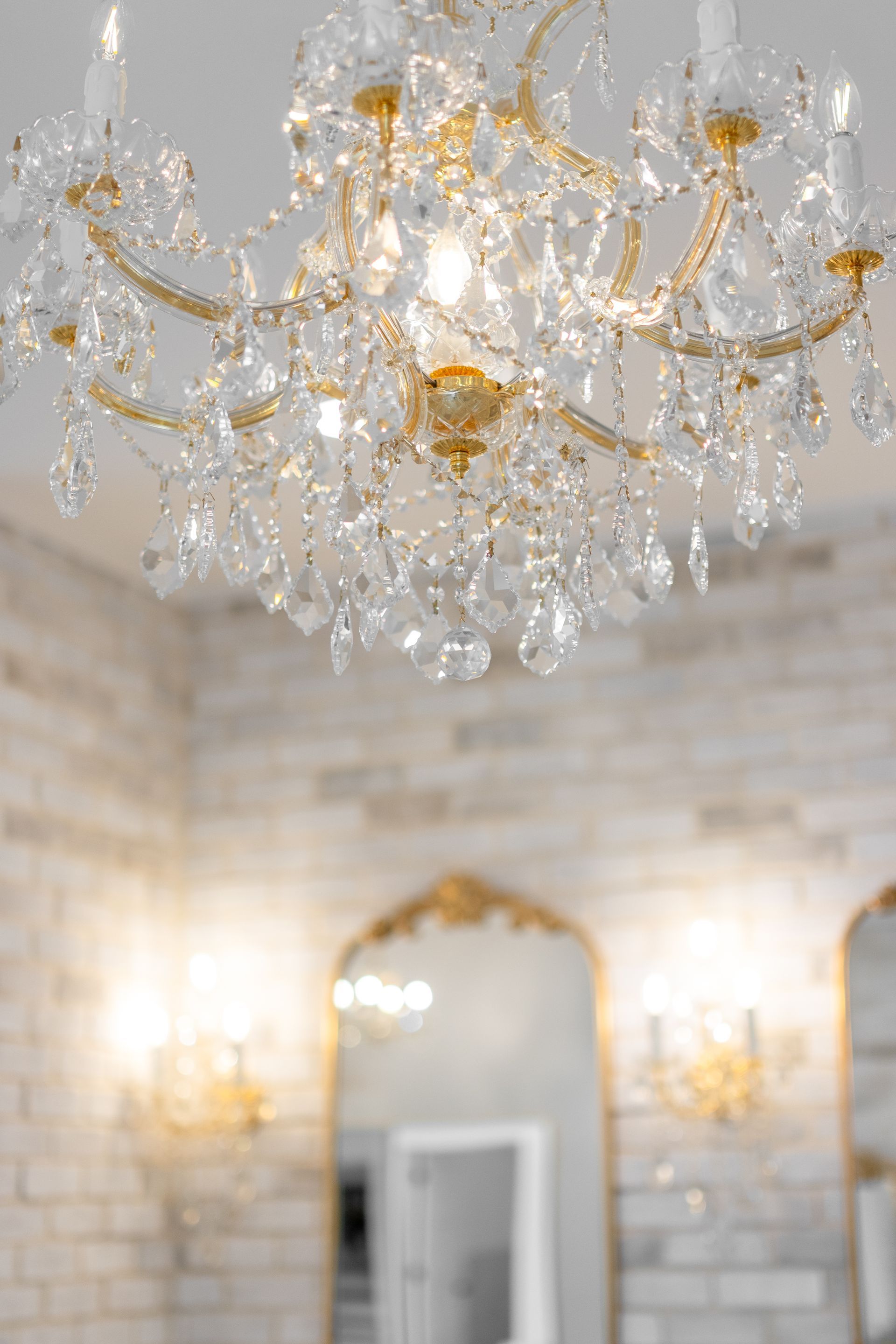 Elegant crystal chandelier in a white brick room with a gold-framed mirror and wall sconces.