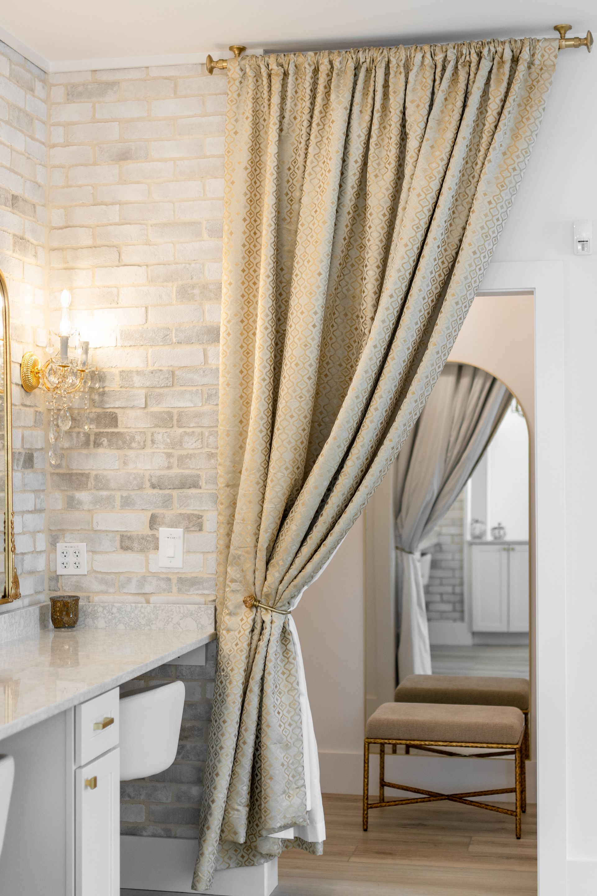 Elegant bathroom with beige drapes, white vanity, and a gold wall sconce beside a mirrored chair.