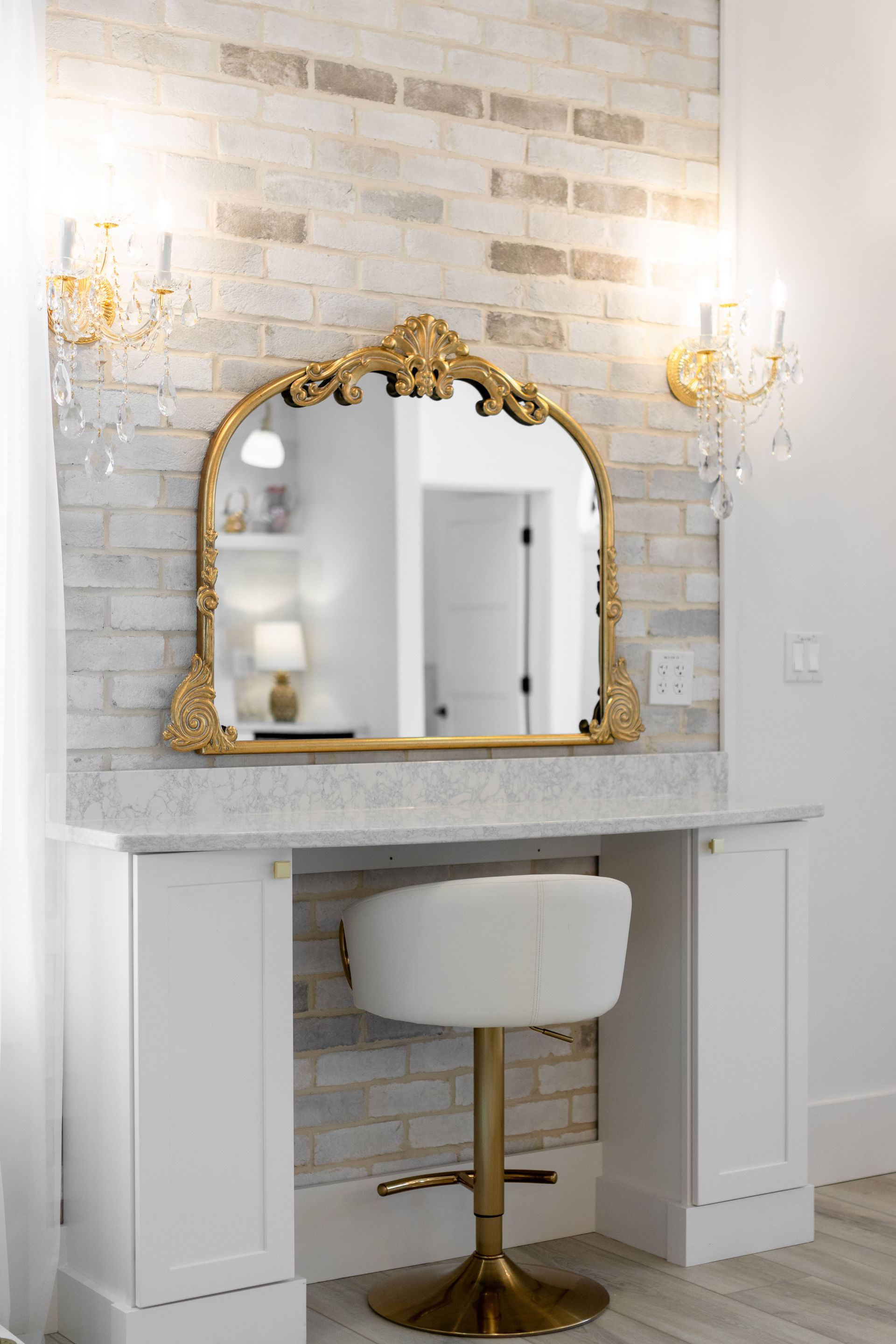 Elegant vanity area with gold-framed mirror, wall sconces, and white tufted stool under a marble counter