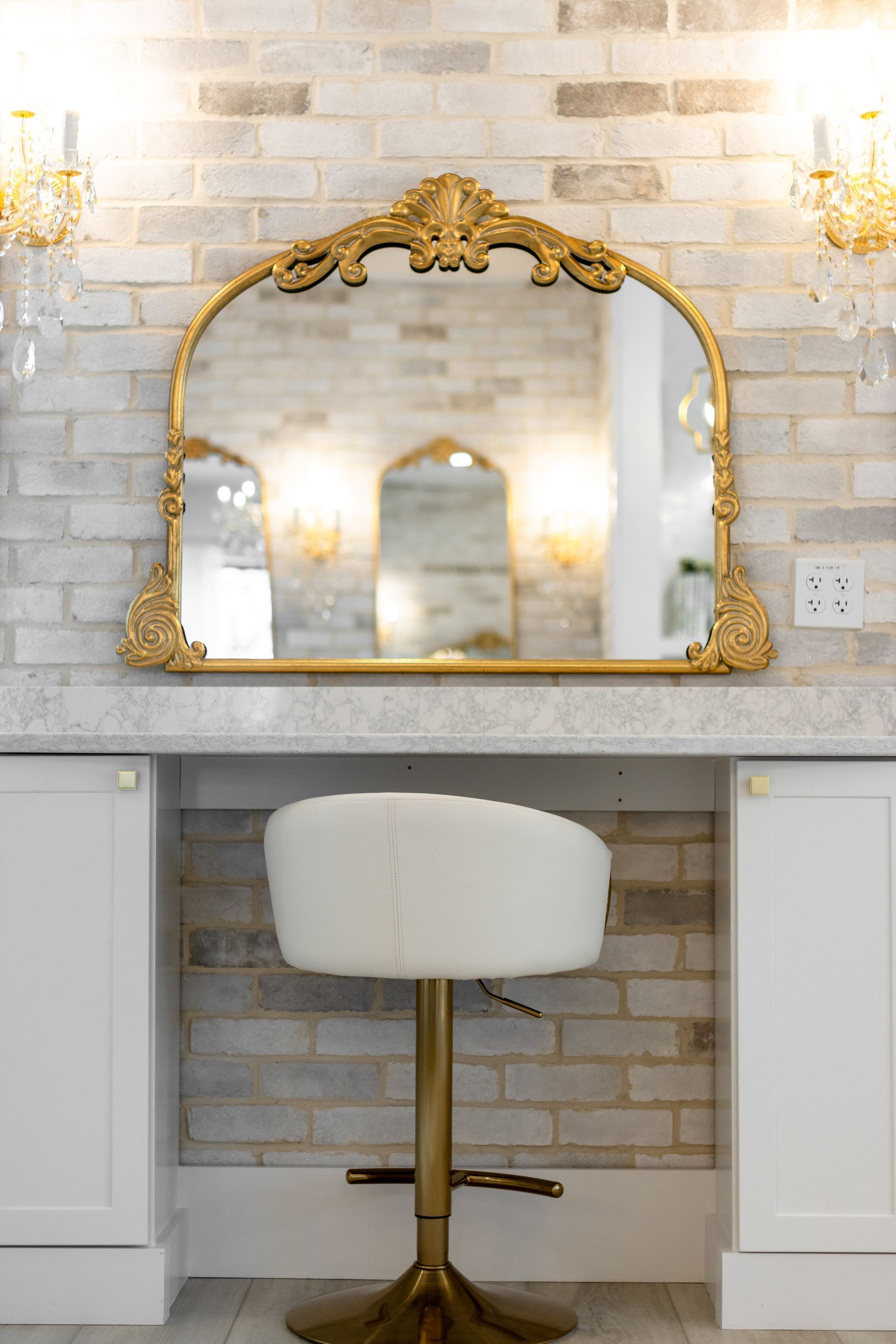 Gold-framed wall mirror above a white vanity with a cushioned stool in a bright bathroom.