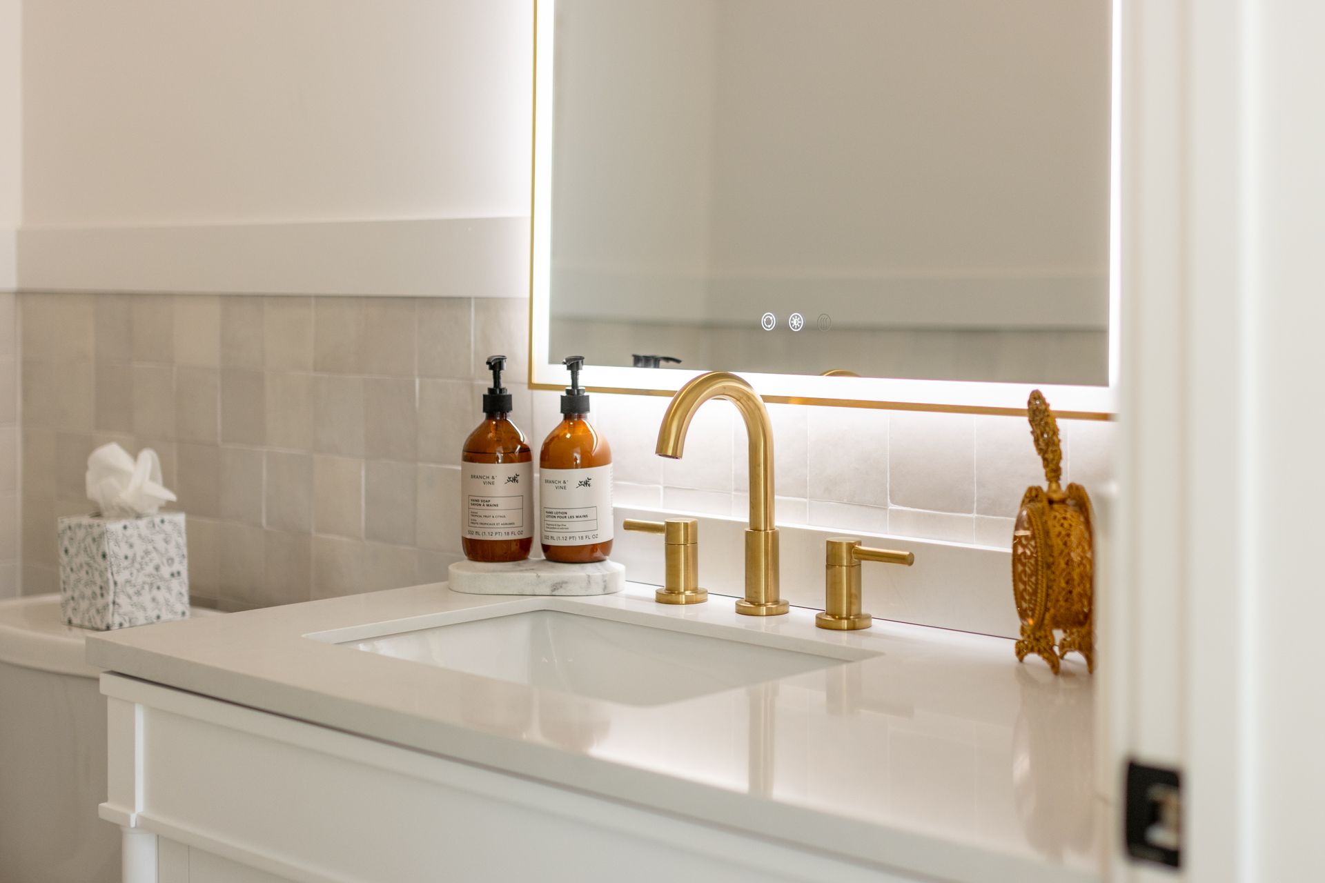 Bright bathroom vanity with white sink, gold faucet, amber soap bottles, and a candle beside a window shade