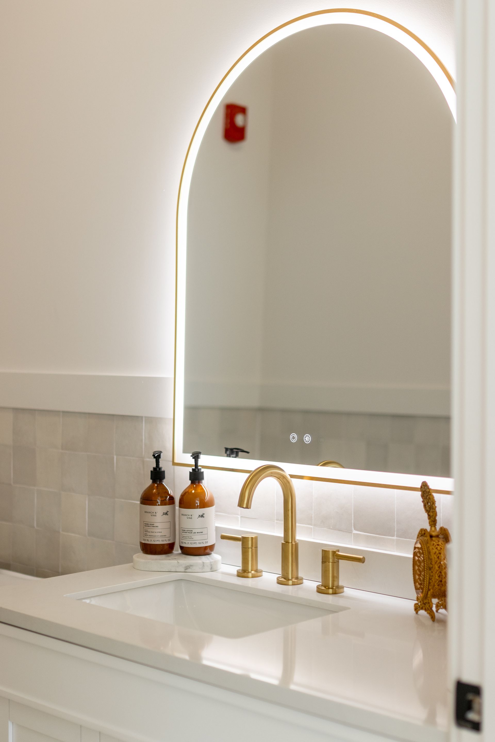 Modern bathroom vanity with arched backlit mirror, gold faucet, white sink, and soap bottles