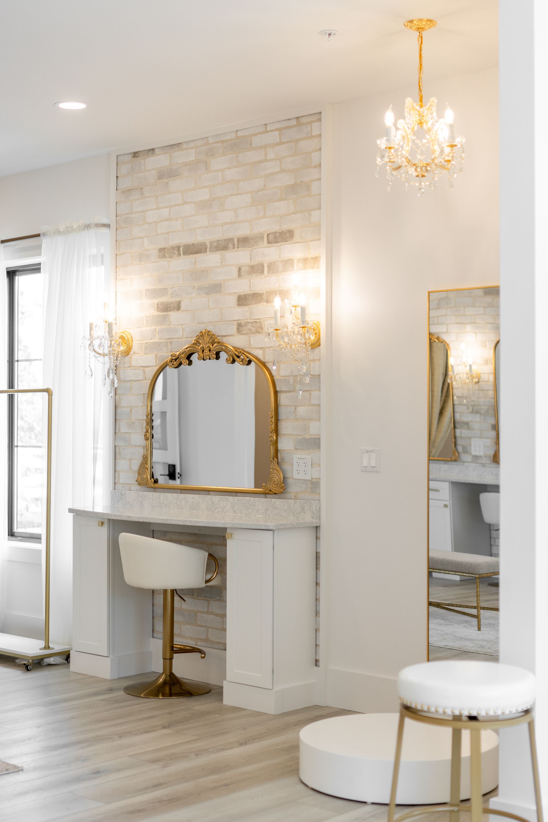 Elegant white vanity area with gold-framed mirror, chandelier, and modern seating in a bright room