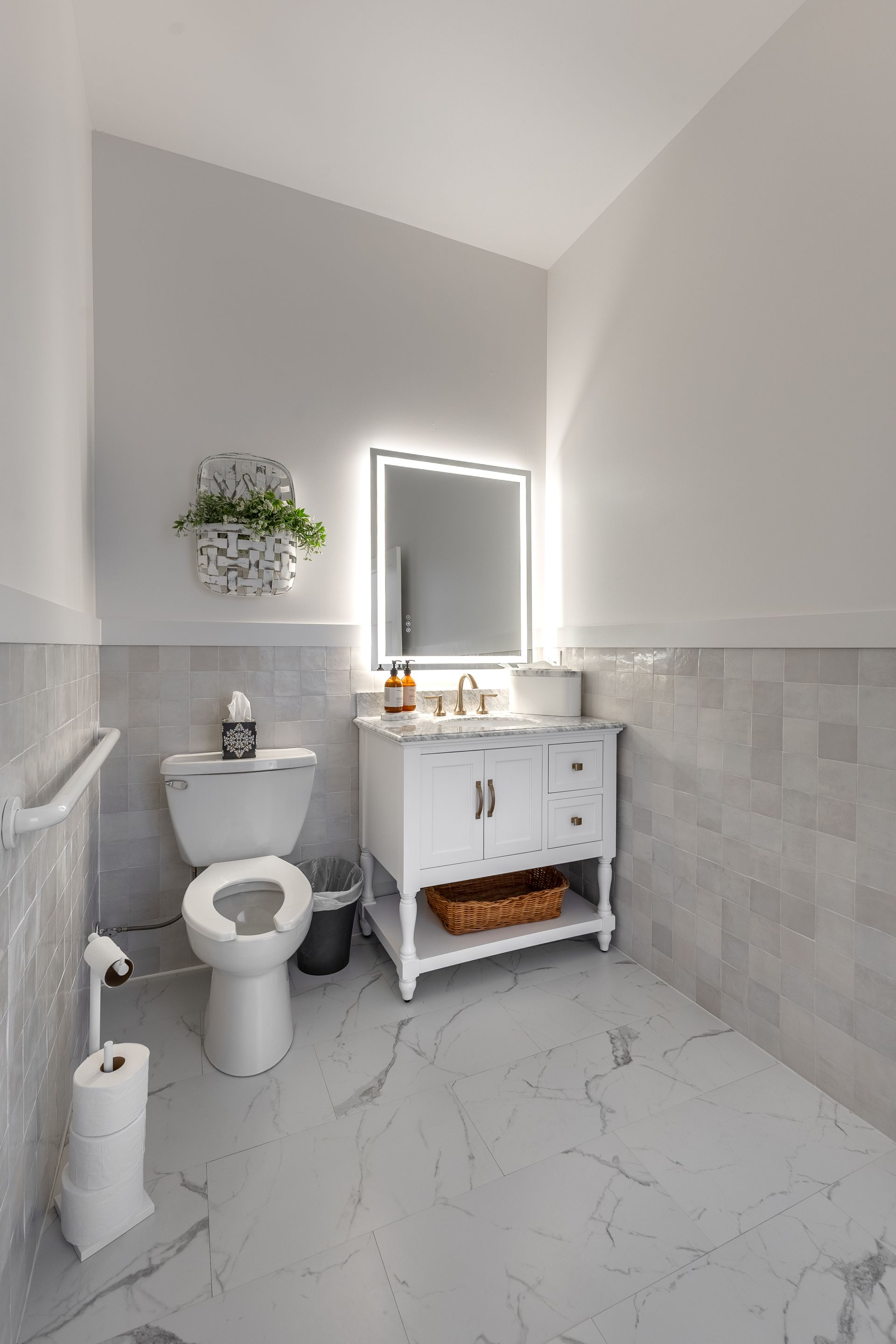 Bright white bathroom with toilet, vanity sink, mirror, wall plant, and marble-patterned tile walls.