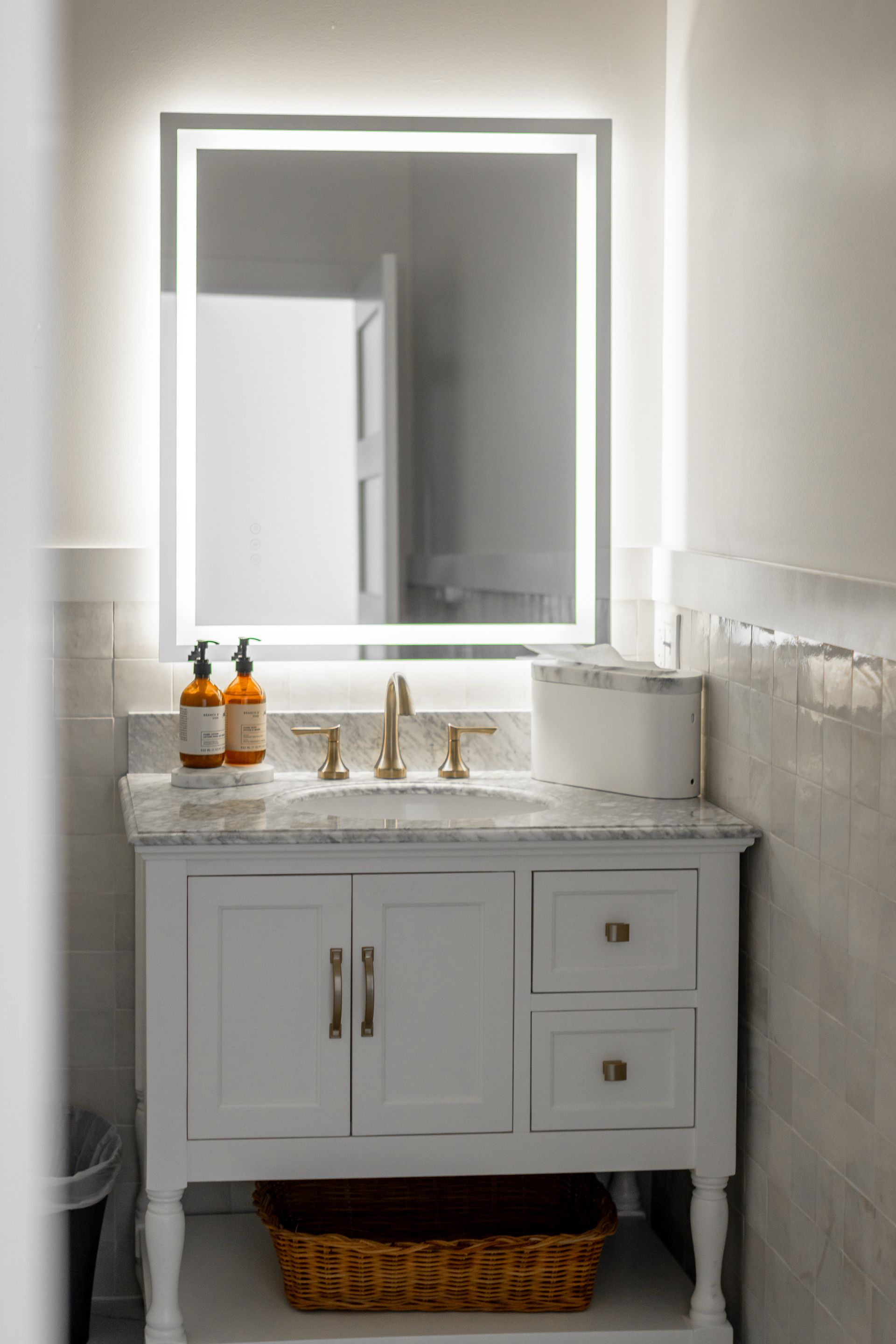 Modern bathroom vanity with lit mirror, white sink cabinet, gold faucet, and wicker basket underneath