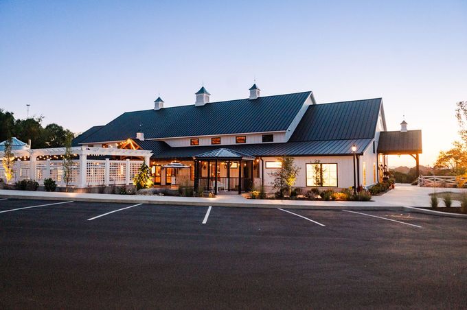 Modern white barn-style building with a dark roof and a parking lot. Dusk lighting.