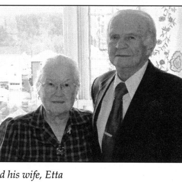 An older couple stands together smiling, next to a window. The man is wearing a suit and the woman a patterned blouse.