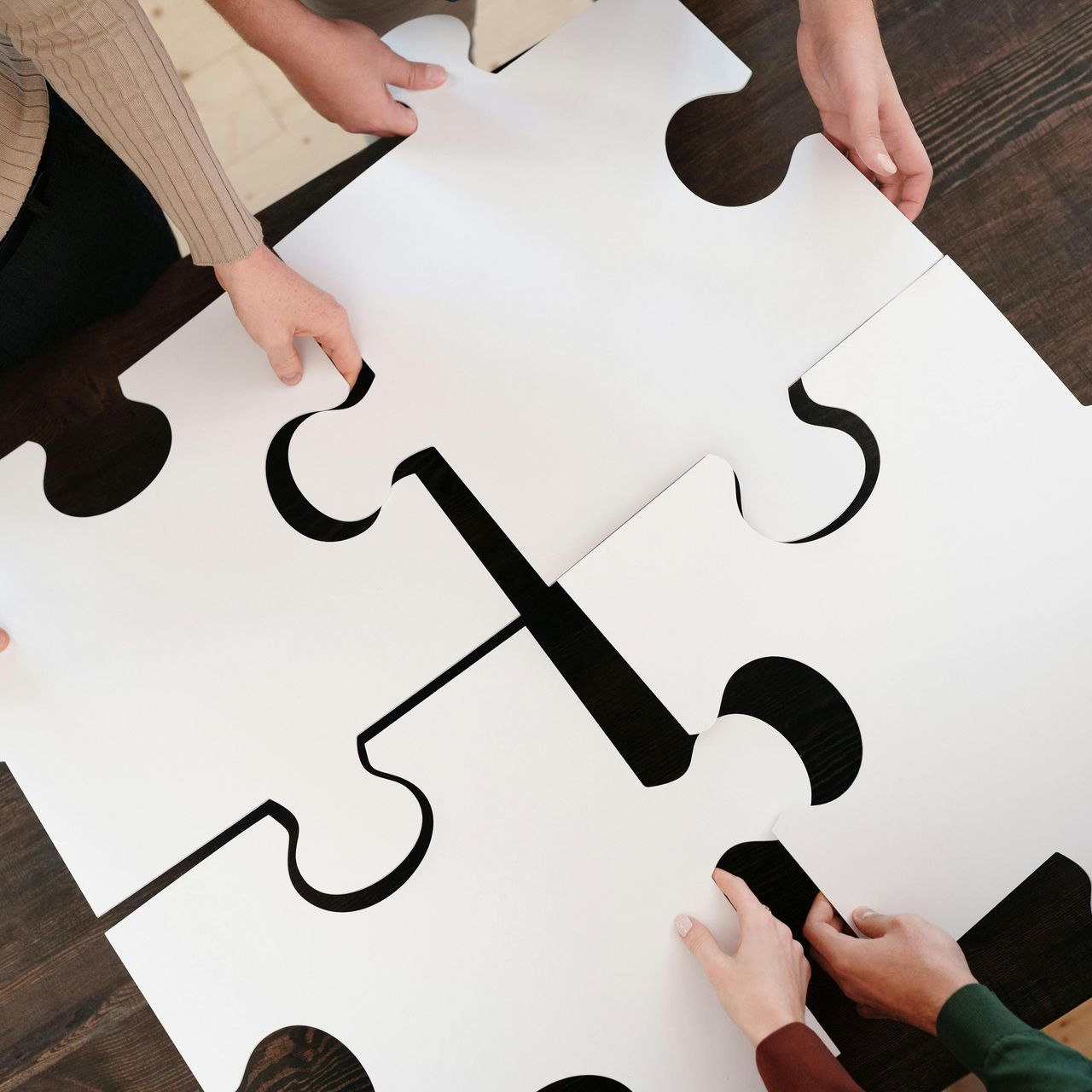 Hände fügen vier große, weiße Puzzleteile auf einer dunklen Holzoberfläche zusammen.