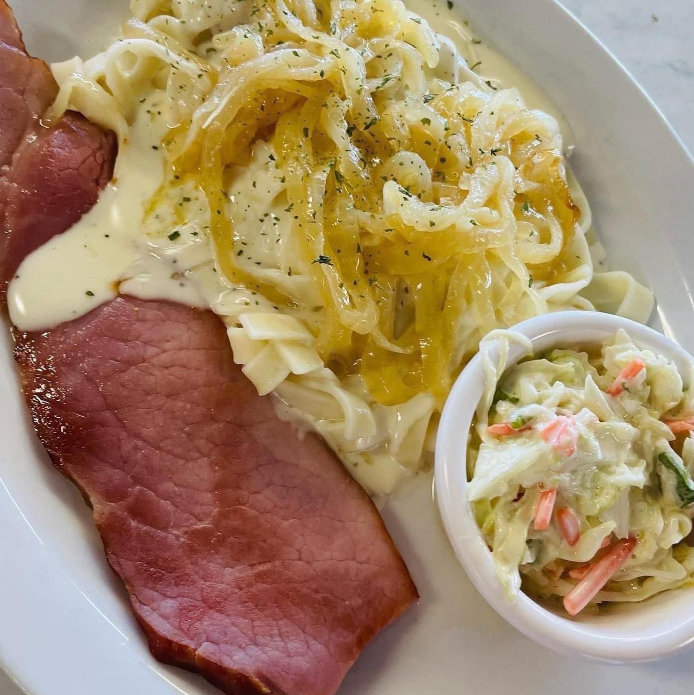 Plate with sliced roast beef, creamy noodles, and a small cup of coleslaw on a white table.