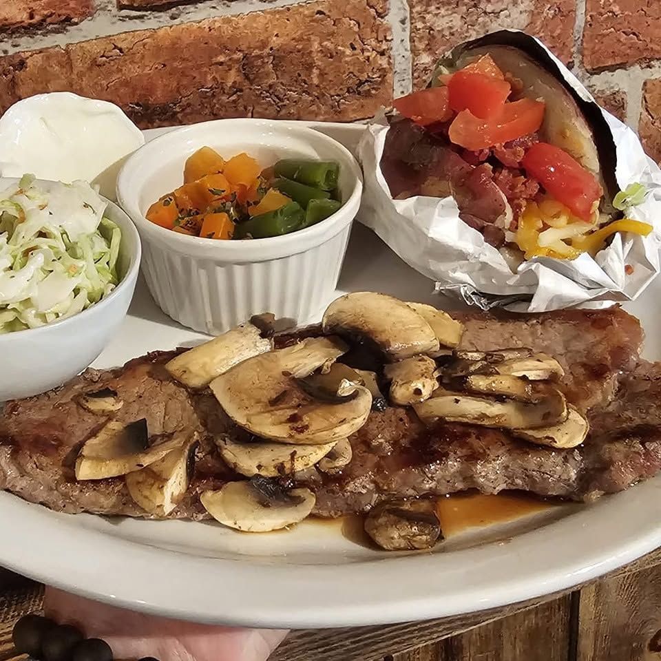 Plated steak topped with mushrooms, with coleslaw, vegetables, mashed potatoes, and a foil-wrapped baked potato