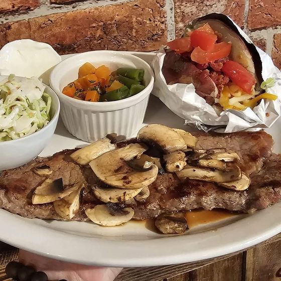 Plated steak topped with mushrooms, with coleslaw, vegetables, and a foil potato on a table.
